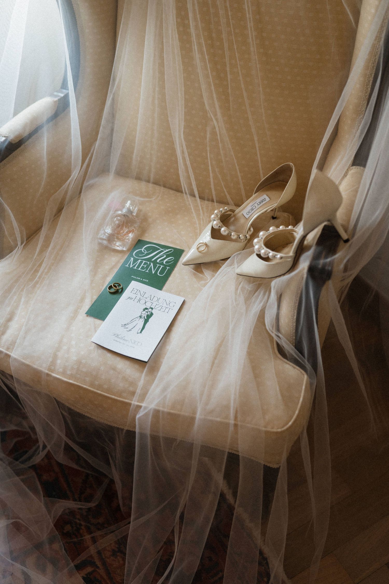 Bridal wedding details flat lay with pearl Jimmy Choo heels, veil, perfume, and menu card on elegant chair.