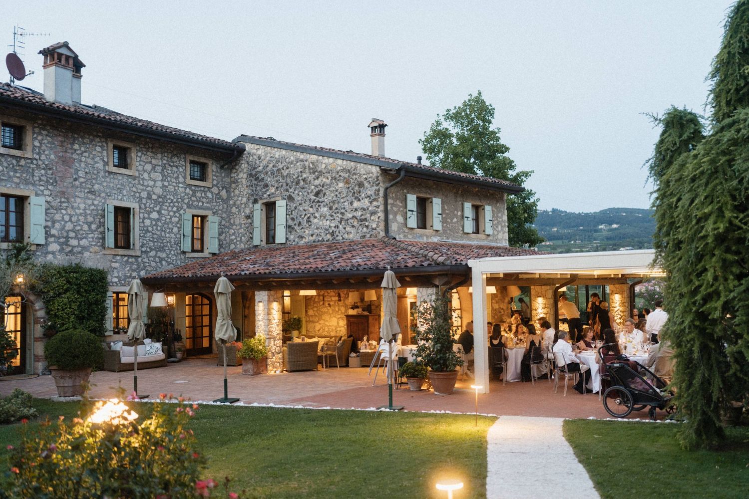 Elegant stone villa with terrace dining at dusk, guests gathered under pergola with warm ambient lighting.