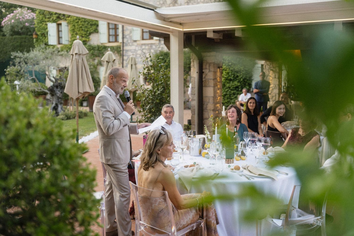 Elegant outdoor wedding reception with guests seated at decorated tables while a man gives a speech under a pergola.