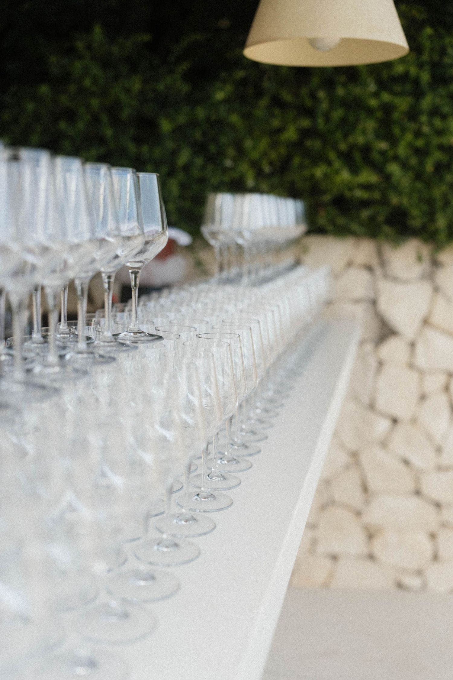 Rows of empty crystal wine and champagne glasses arranged on a white bar table at an elegant outdoor event.