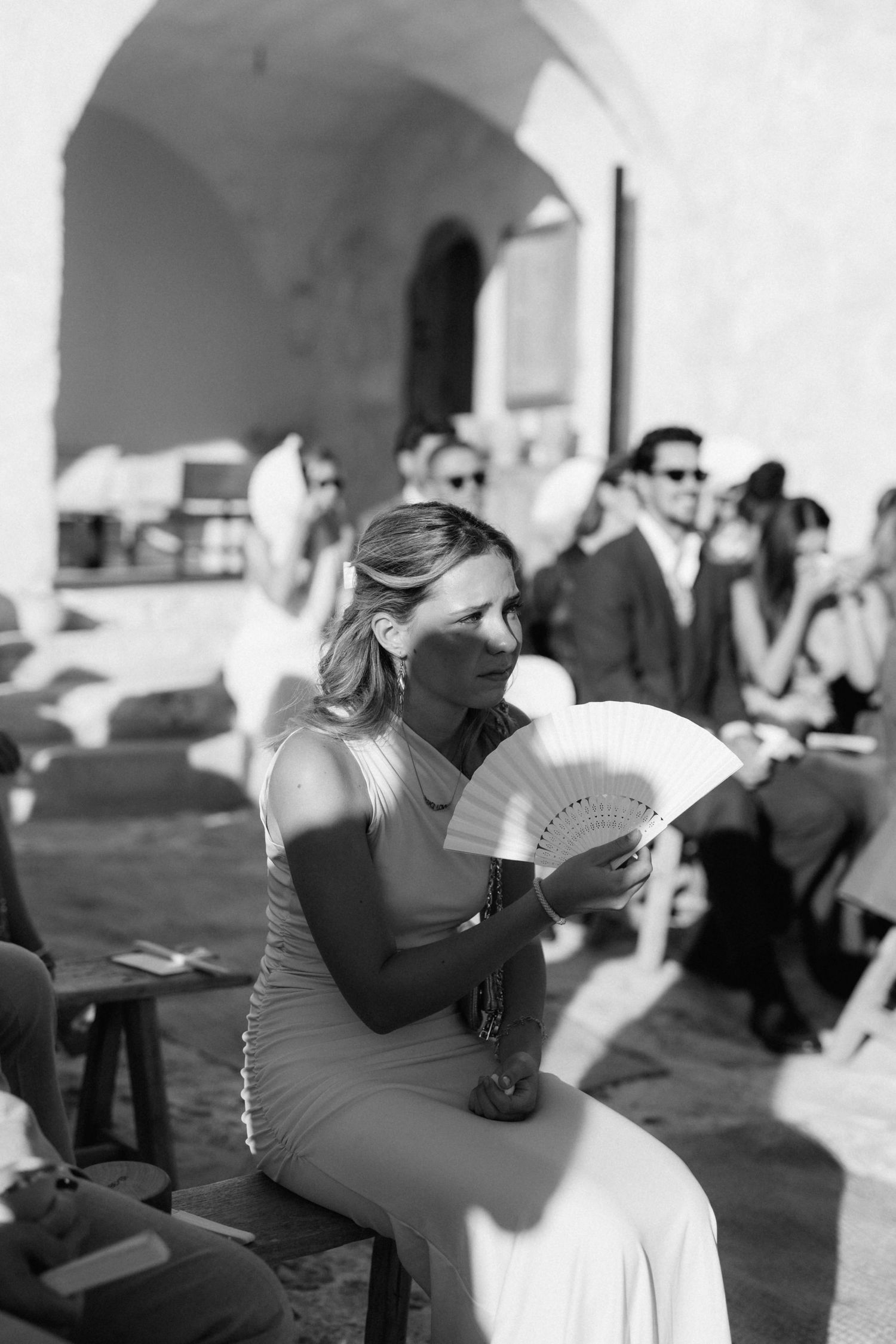 Black and white photo of a woman in a fitted dress holding a white hand fan while seated among guests at an outdoor wedding.