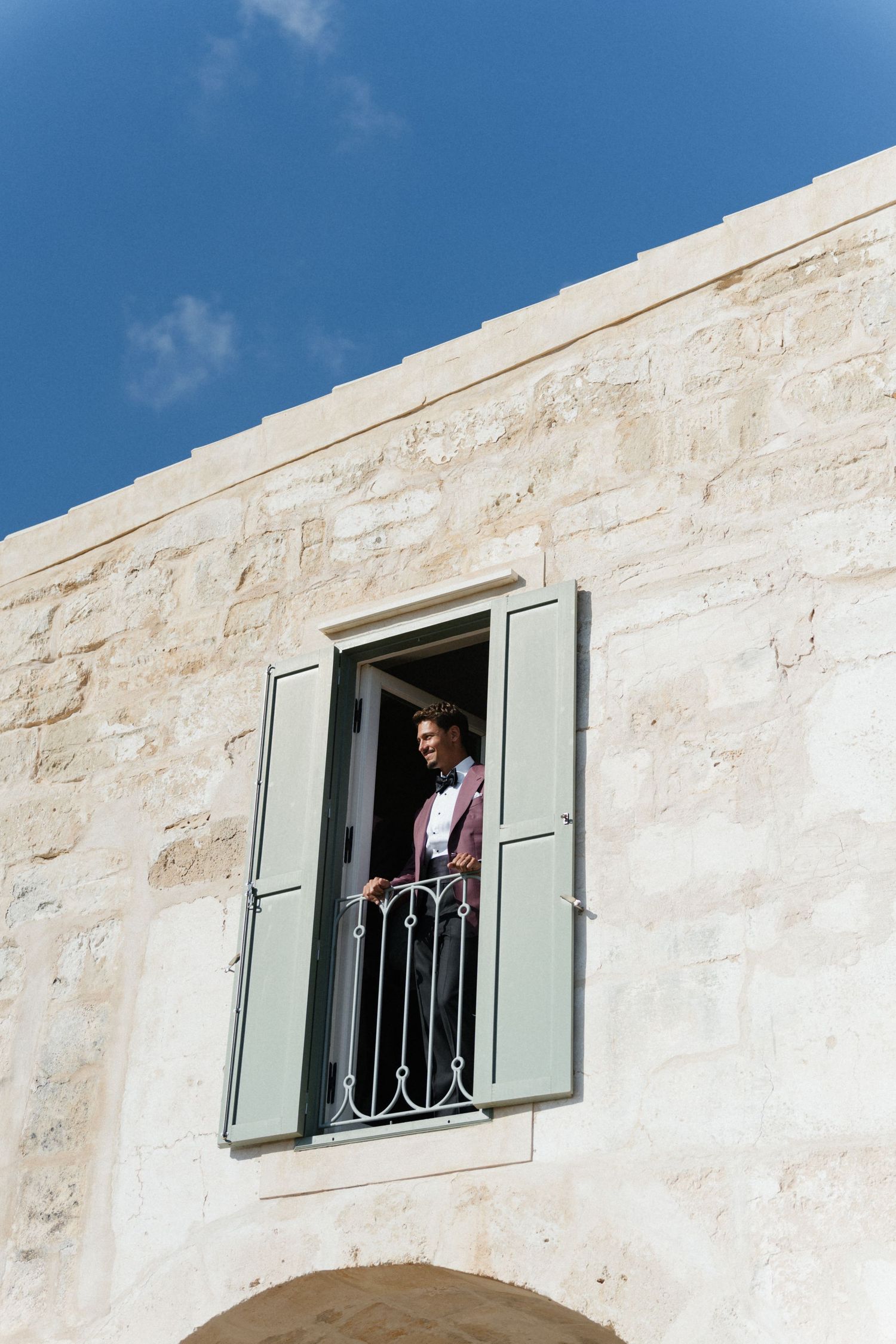 Groom in a suit stands on a wrought-iron balcony between sage green shutters on a sunlit limestone building.