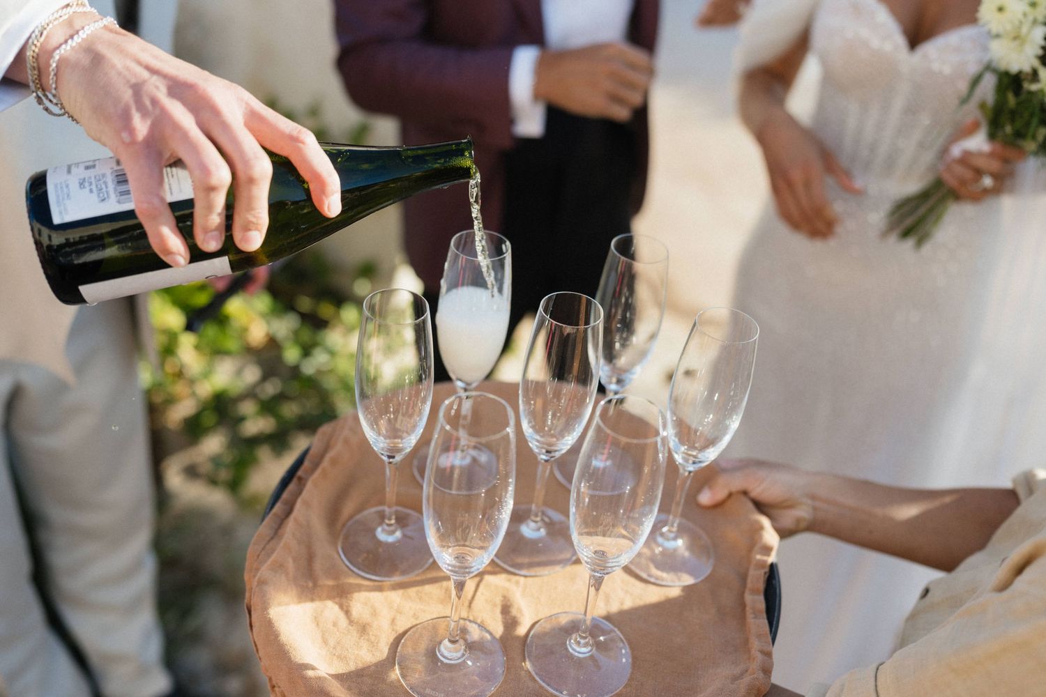 A hand pours champagne into rows of glasses on a table as a bride holding a bouquet stands in the background.