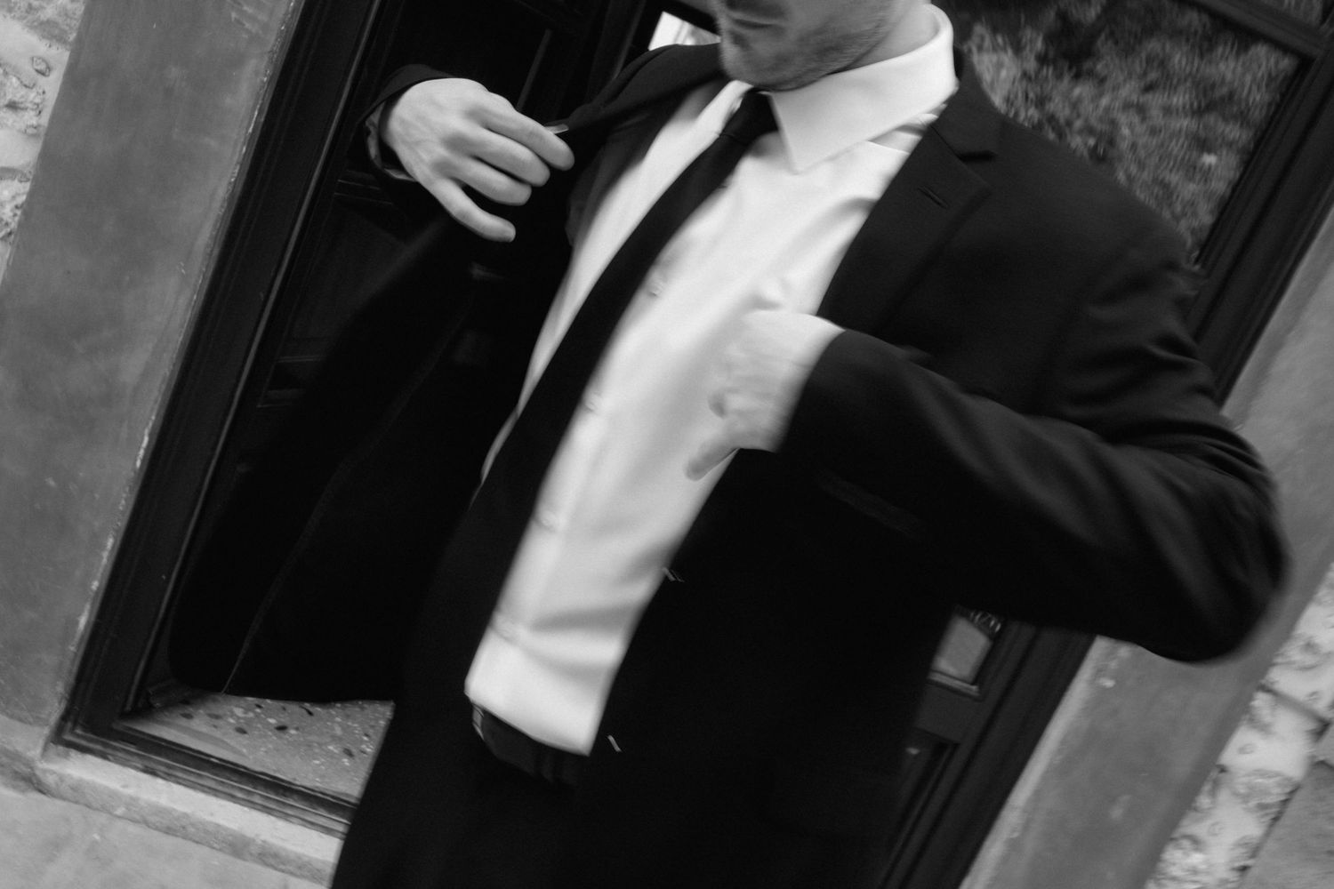 Black and white photo of a man in a dark suit and tie adjusting his jacket beside a doorway.
