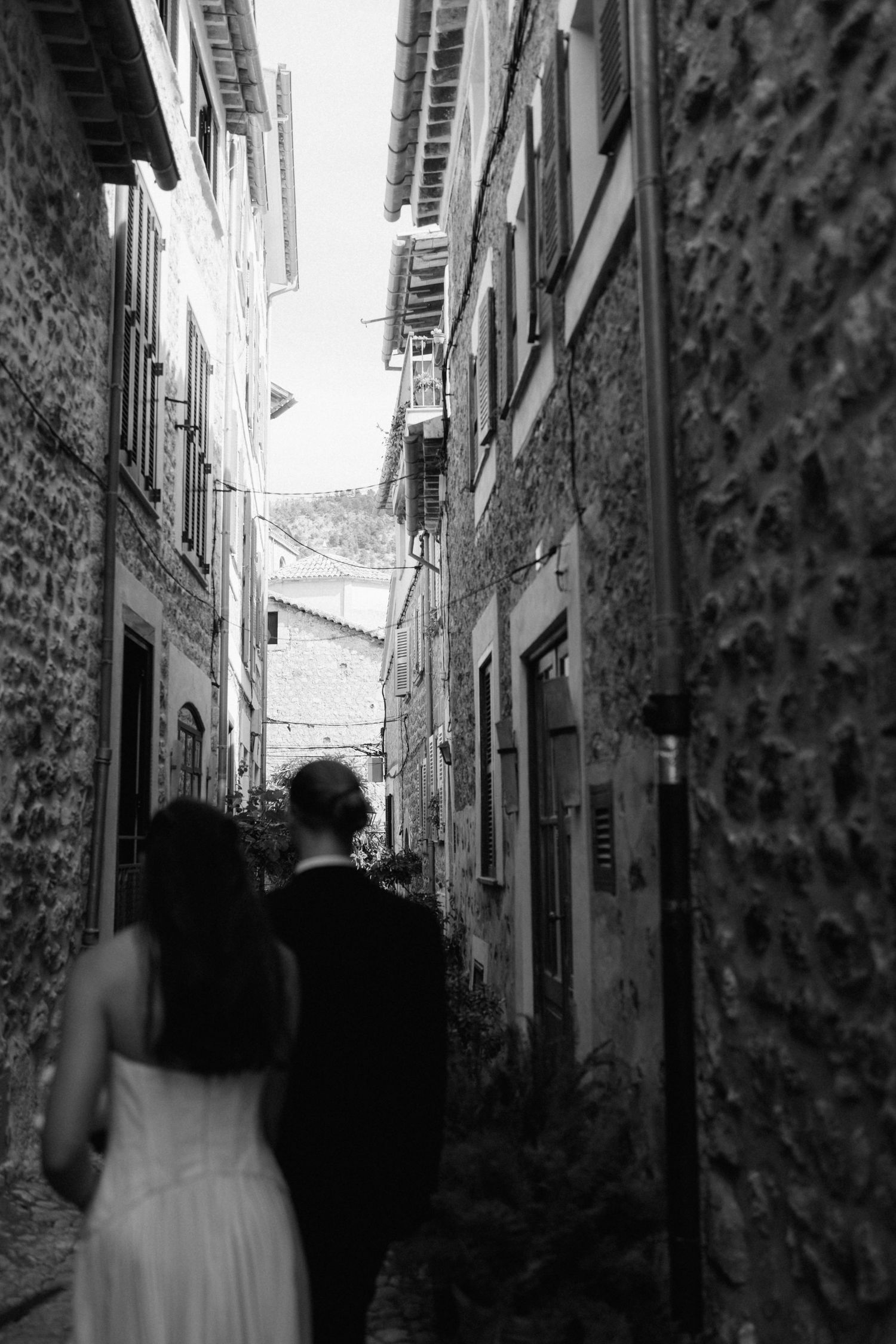 Black and white photo of a bride and groom walking away along a narrow stone alley between old village buildings.