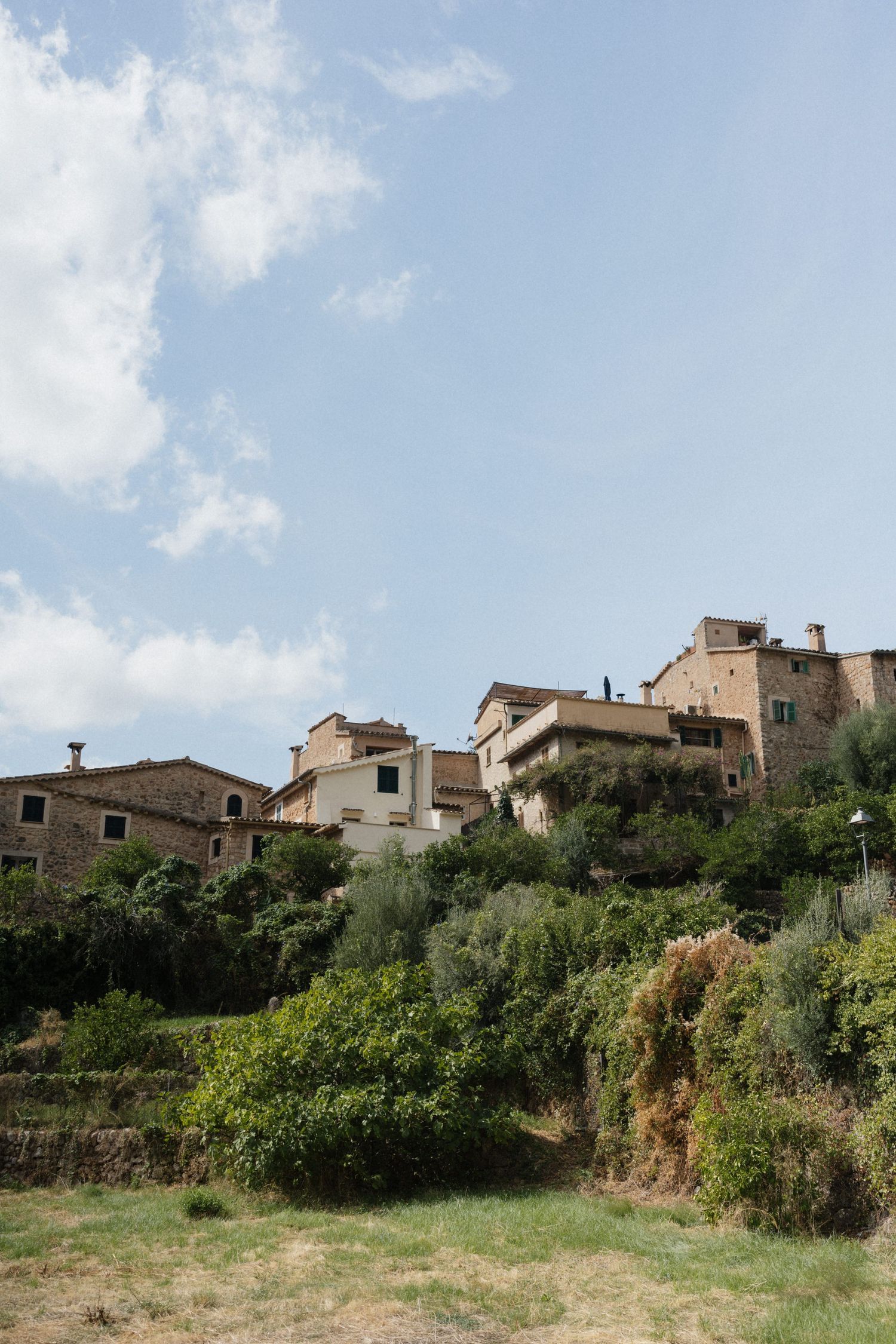 Stone houses with terracotta roofs sit on a lush green hillside beneath a blue sky with white clouds in a charming Mediterranean village.