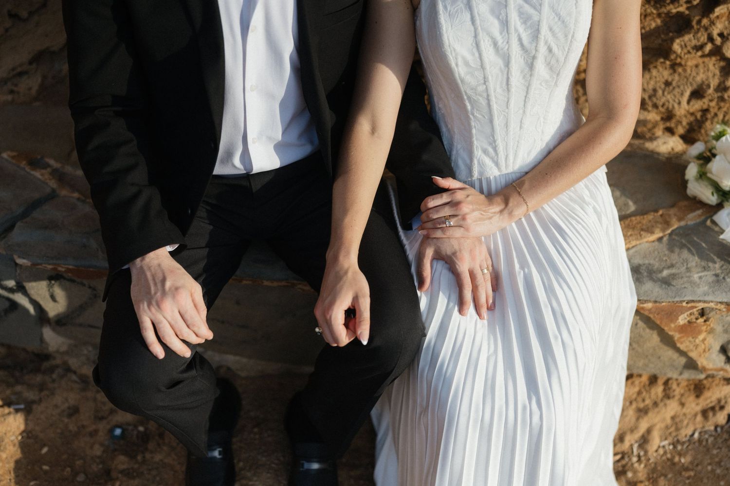 Bride in a white pleated dress and groom in a black suit sit side by side on a rock, holding hands in a close-up wedding moment.