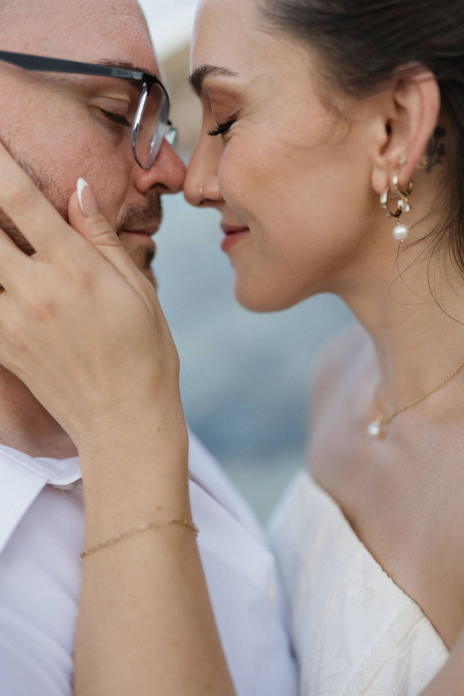 Close-up of a smiling bride in pearl earrings touching noses with her glasses-wearing groom, who gently cradles her hand.