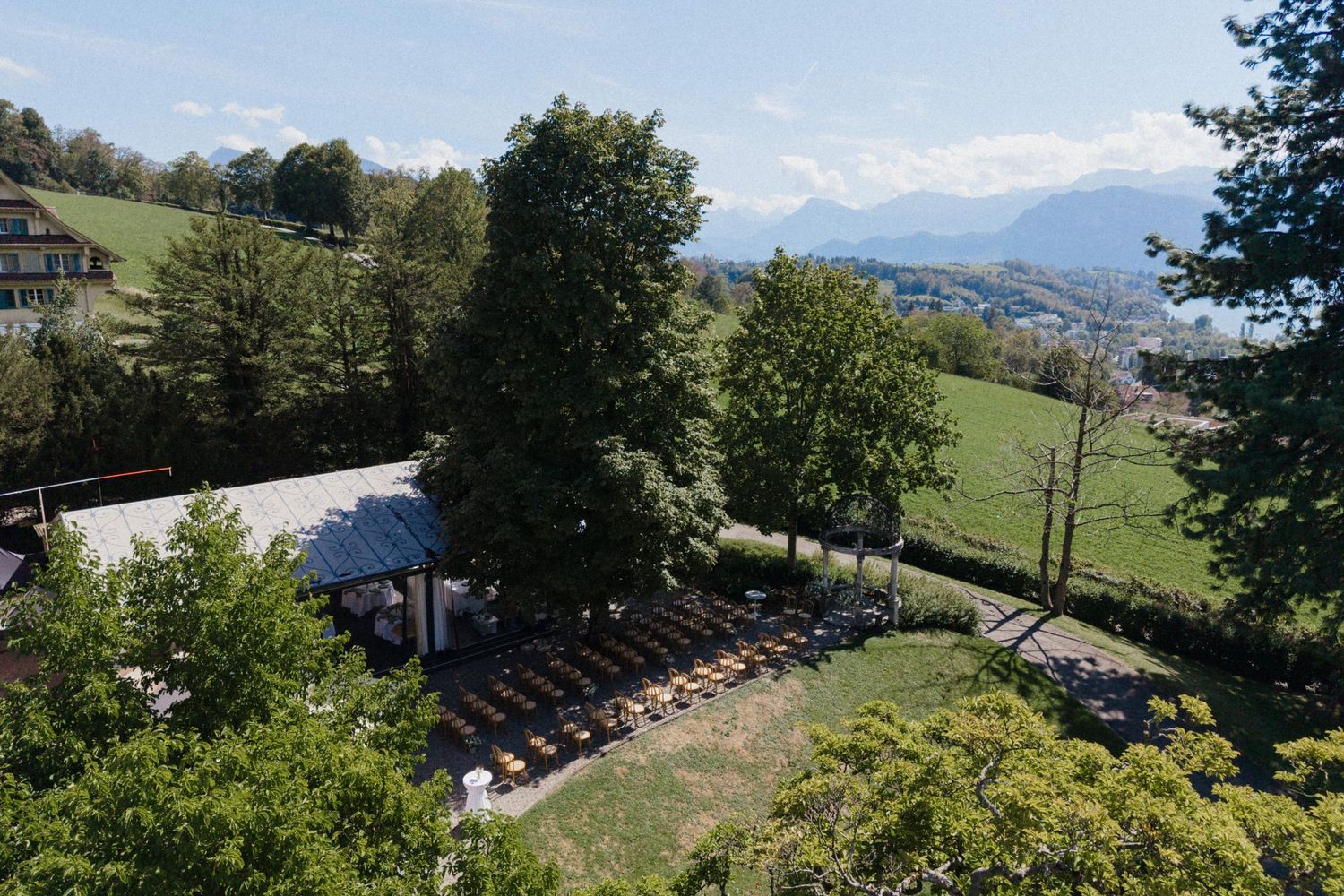 Aerial view of an outdoor event venue with rows of chairs set up on green lawn, surrounded by trees and Alpine mountains.