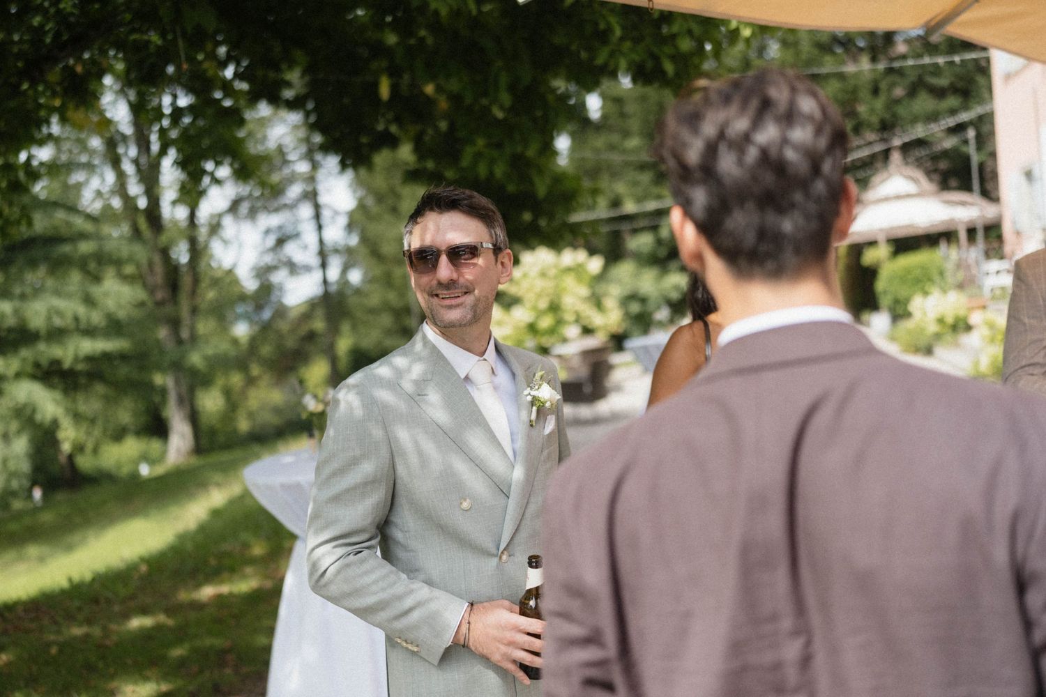 Smiling groom in a light grey suit wearing sunglasses holding a champagne bottle at an outdoor wedding reception.