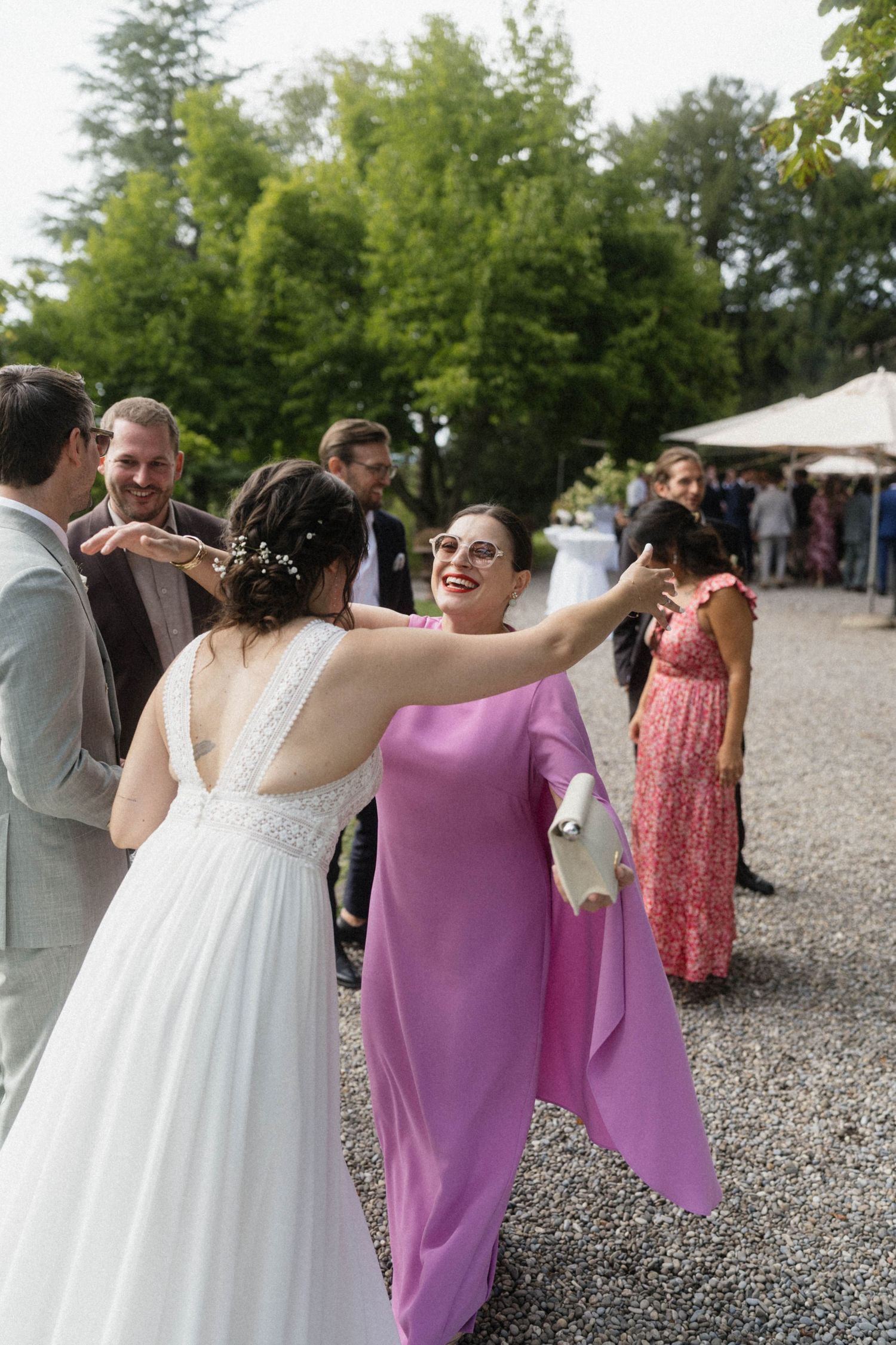 Bride in white gown embracing a guest in purple dress during outdoor wedding reception on gravel path.