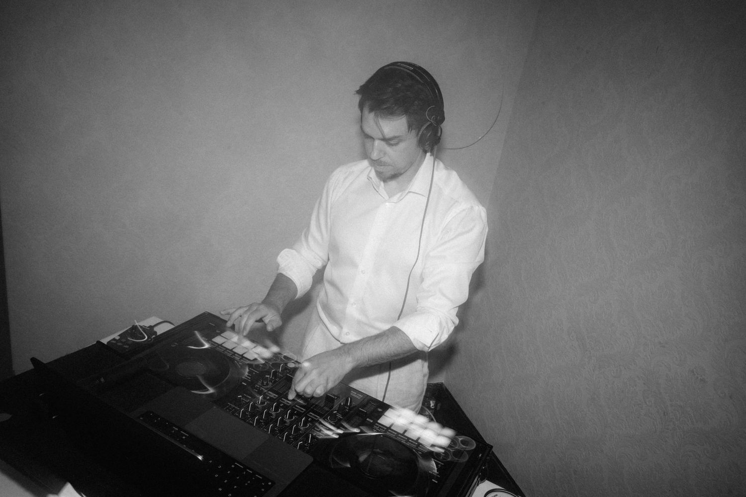 Black and white photo of a DJ wearing headphones working at a mixing console in a dimly lit venue.