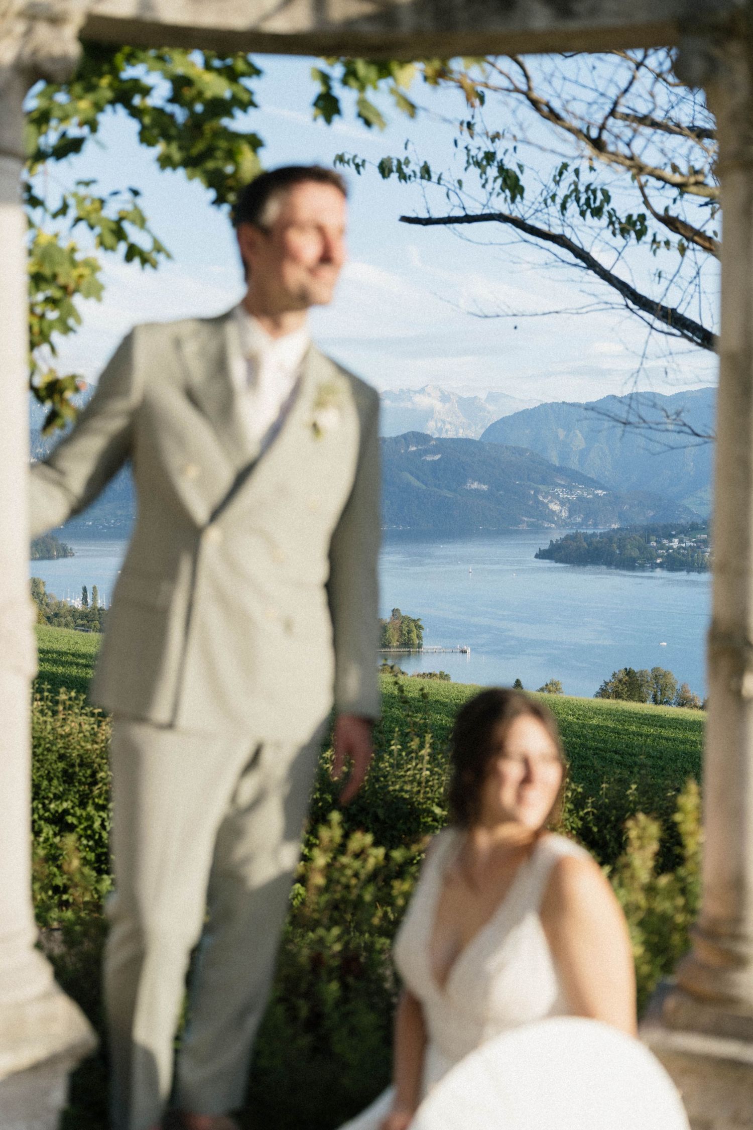 Groom in grey suit stands with bride in white dress framed by columns overlooking a scenic lake.
