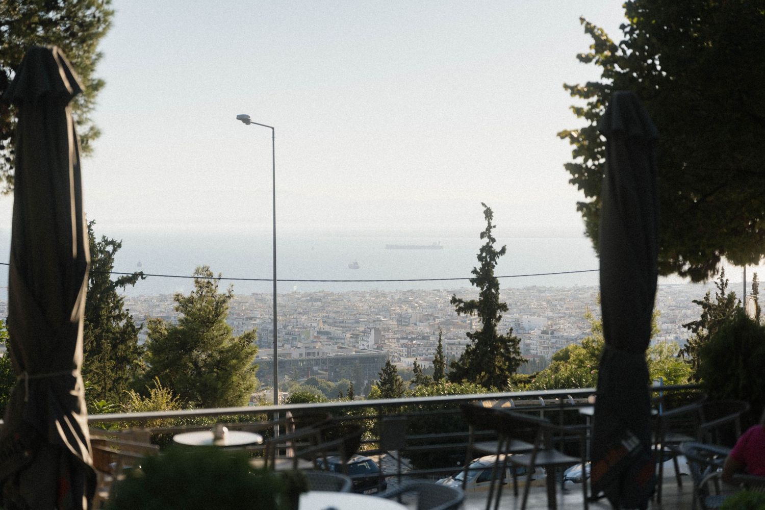 Panoramic view of Thessaloniki city and sea from a hillside cafe terrace with outdoor seating and trees.