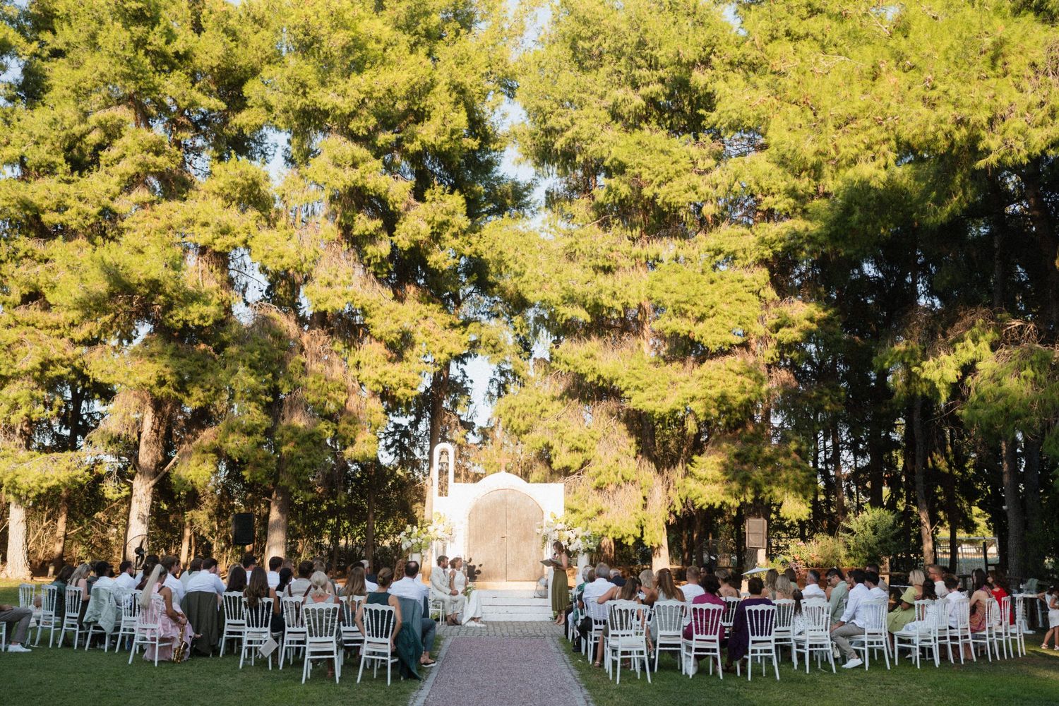 Outdoor wedding ceremony with guests seated on white chairs beneath tall trees leading to an elegant arch backdrop.