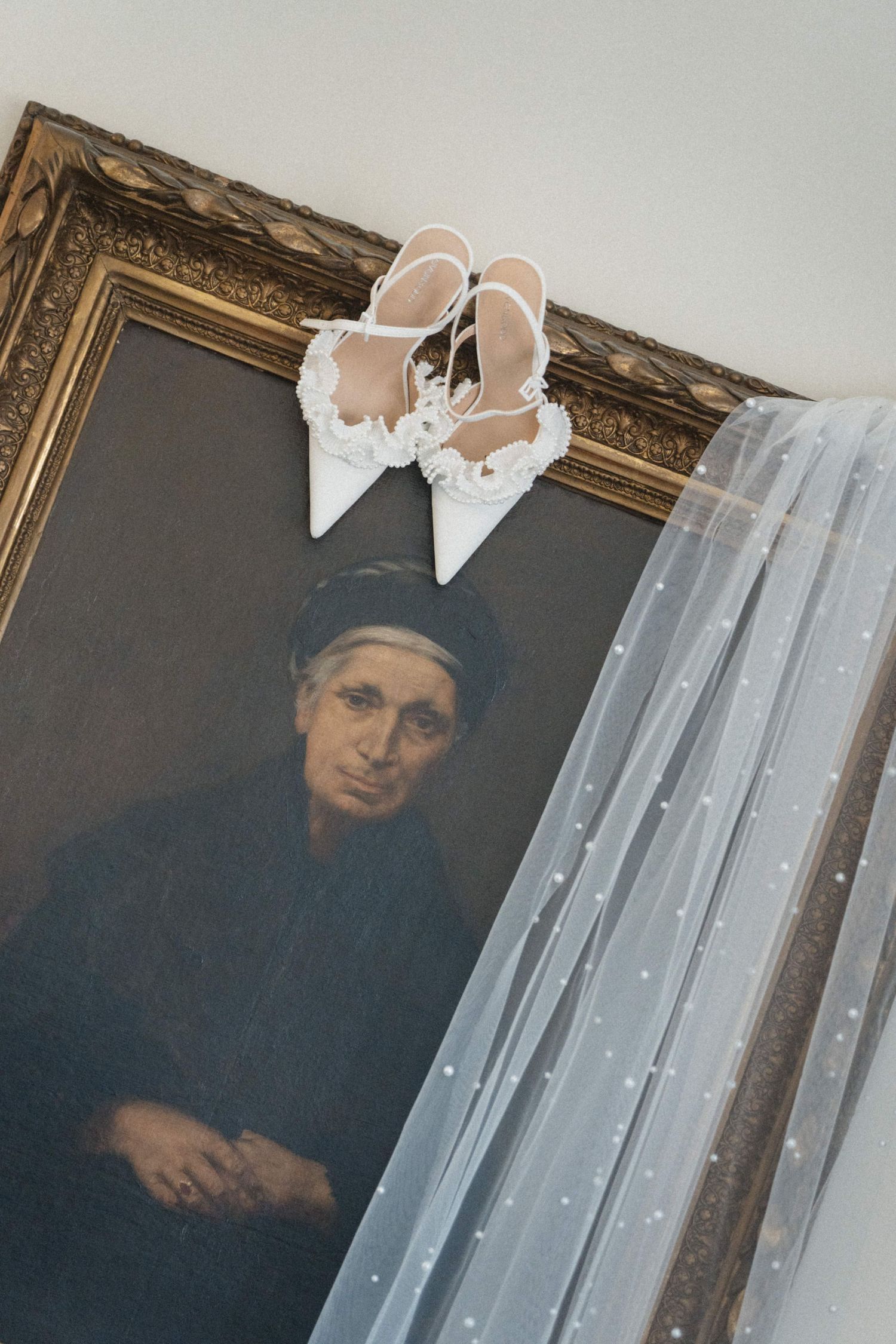 White bridal heels with floral details rest atop a vintage portrait painting beside a polka dot wedding veil.