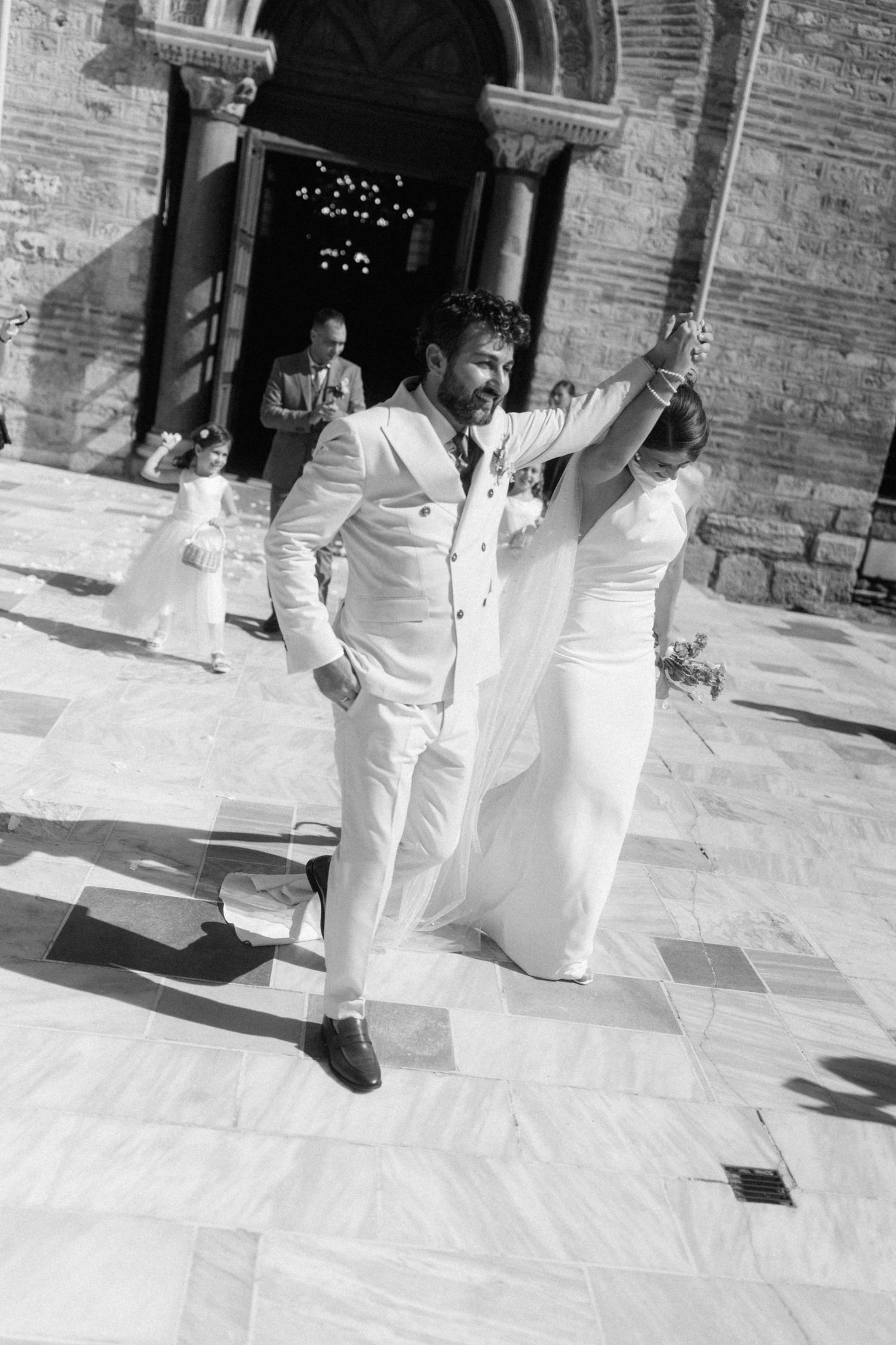 Black and white photo of a joyful newlywed couple exiting a church, bride in gown and groom in white suit.