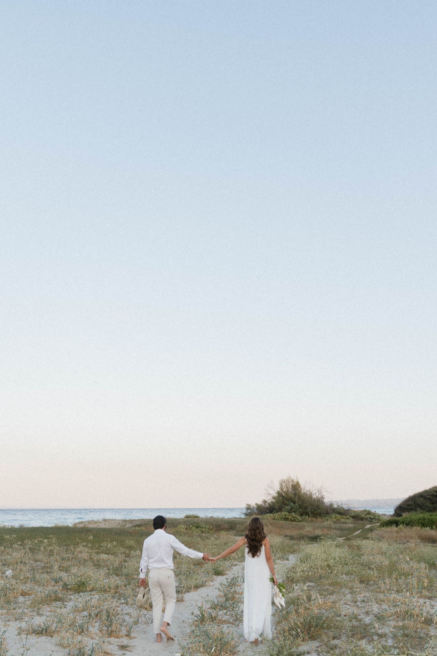 Bride and groom holding hands walking toward ocean on sandy beach path at dusk in white outfits.
