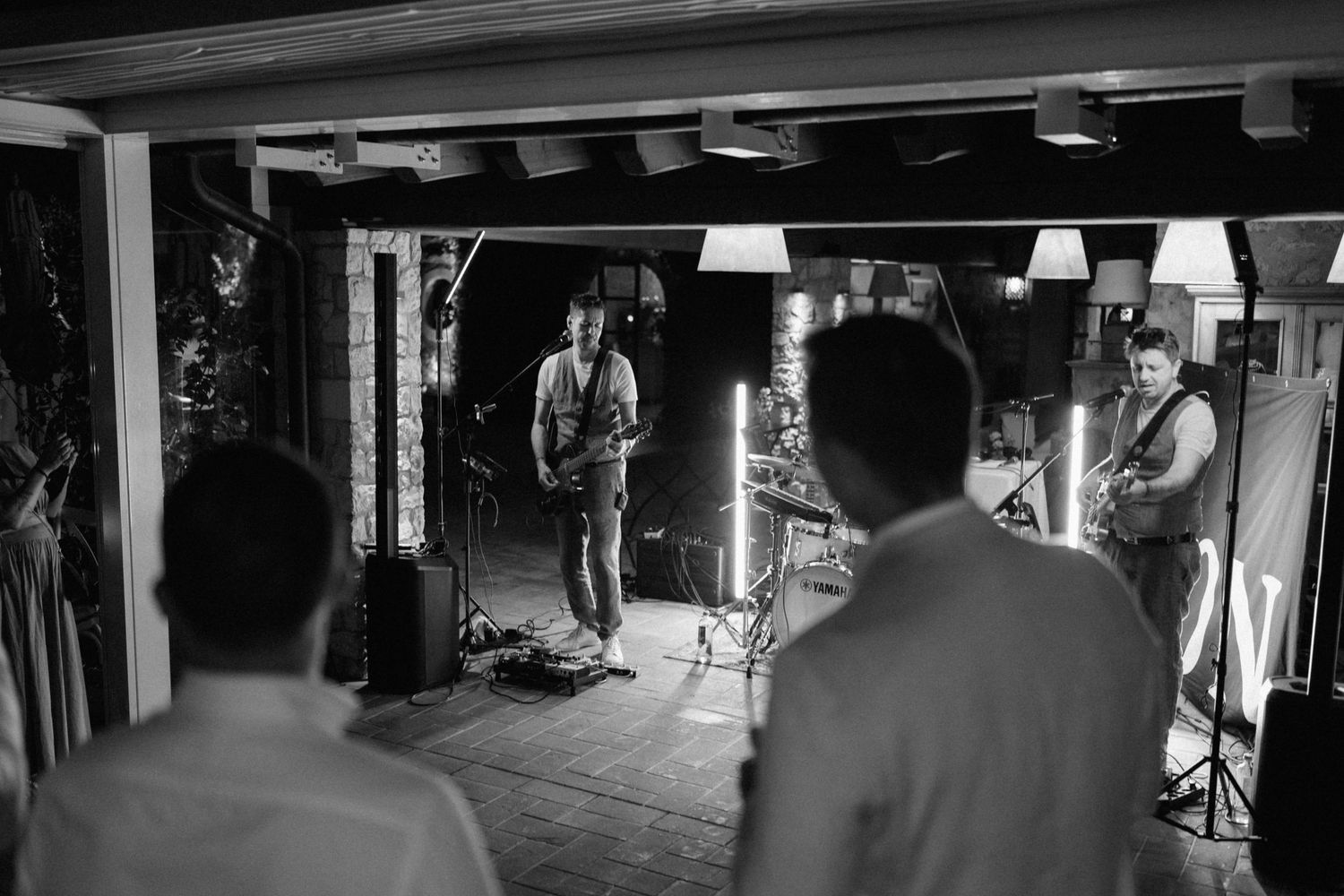 Black and white photo of a live band performing in a rustic venue with studio lighting equipment visible.