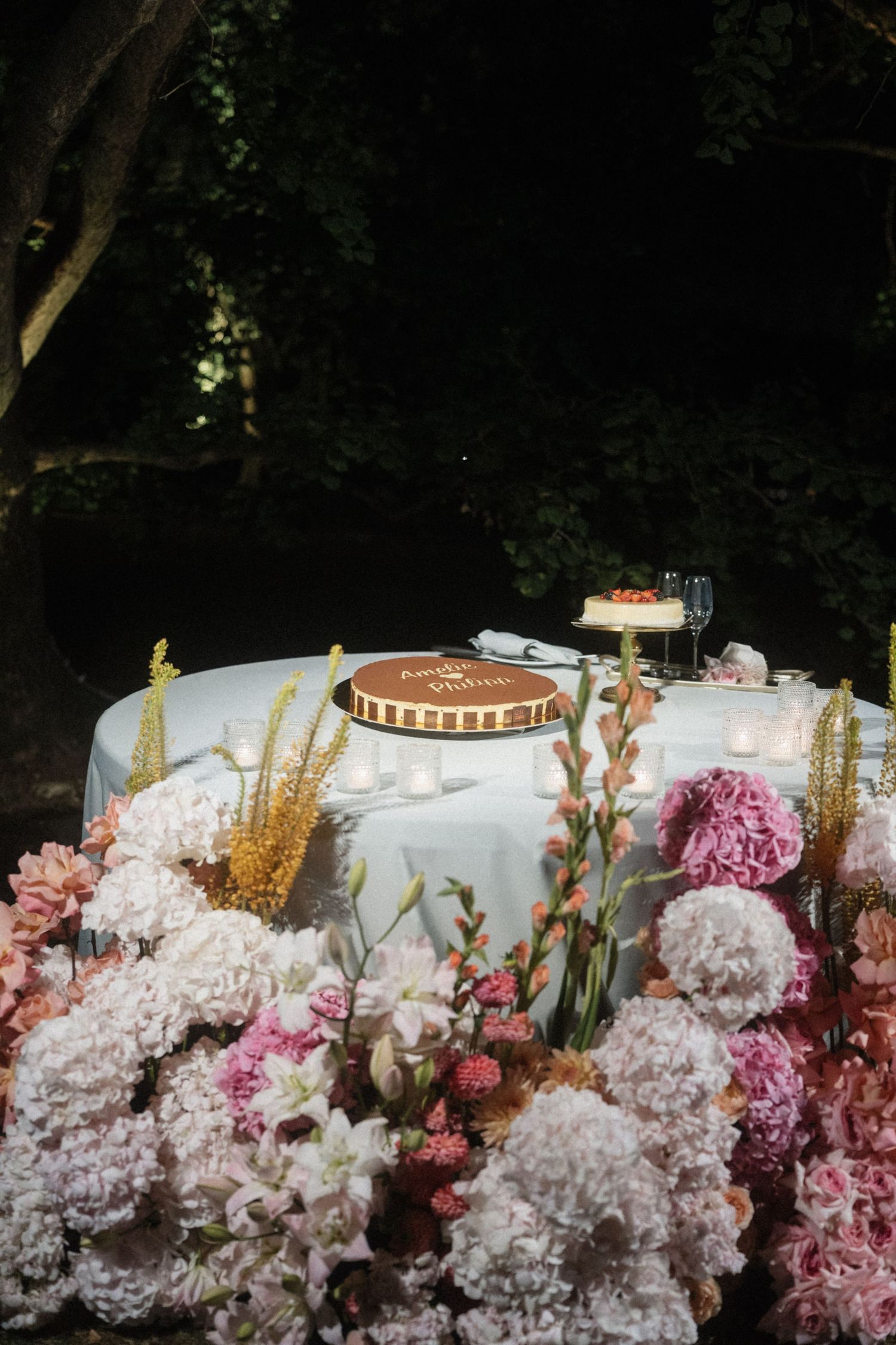 Elegant outdoor dessert table with floral arrangements of pink peonies, candles, and a decorated cake at night.