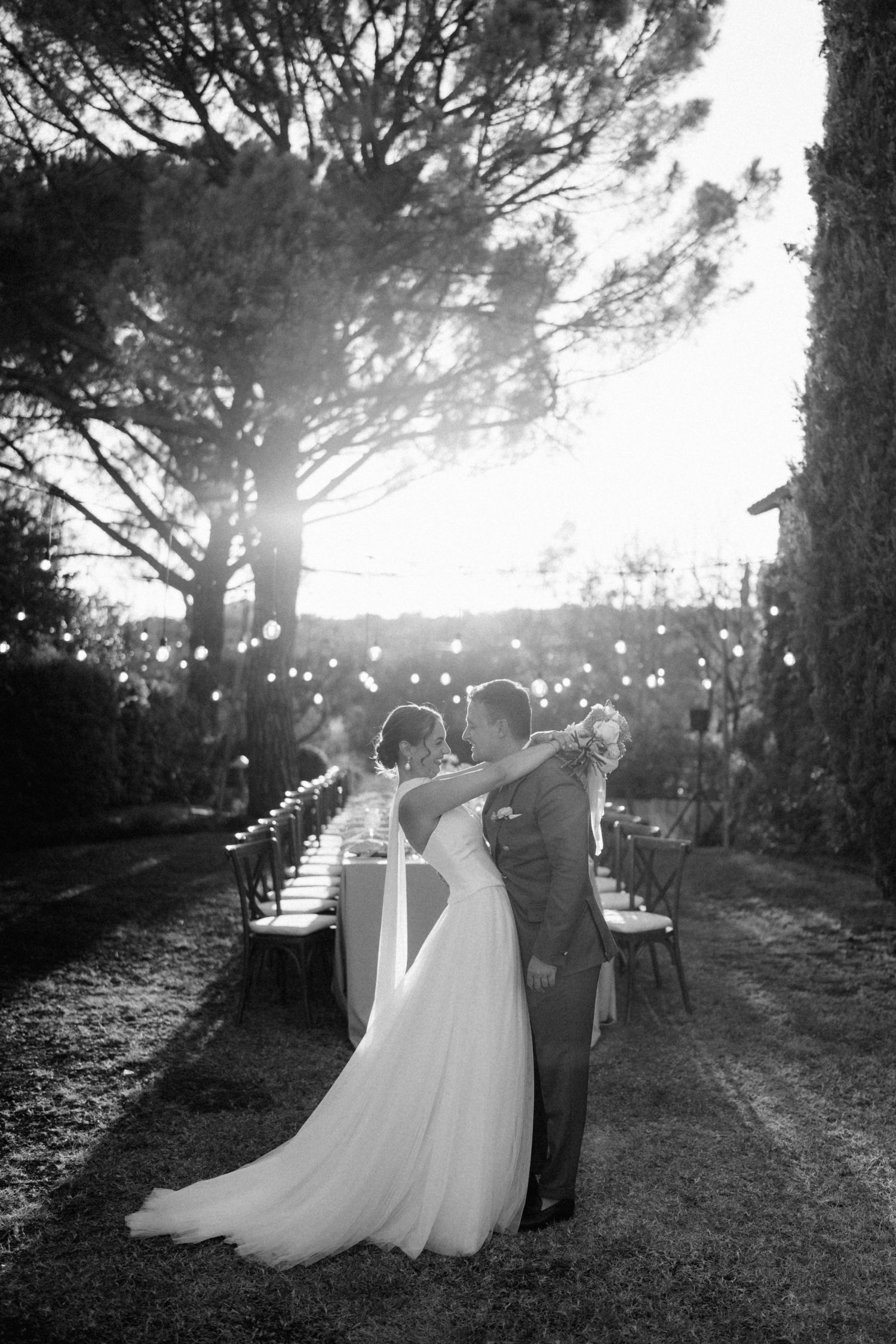 Black and white photo of a bride and groom embracing outdoors at dusk with string lights and a long reception table.