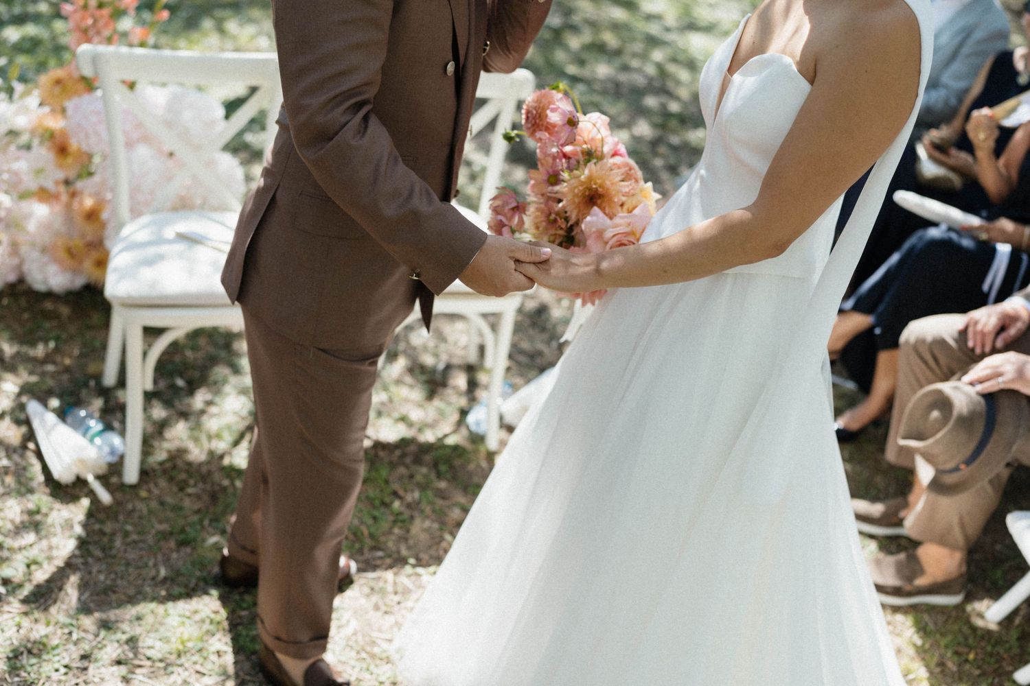 Bride in white gown holding pink bouquet exchanges vows with groom in brown suit at outdoor wedding ceremony.
