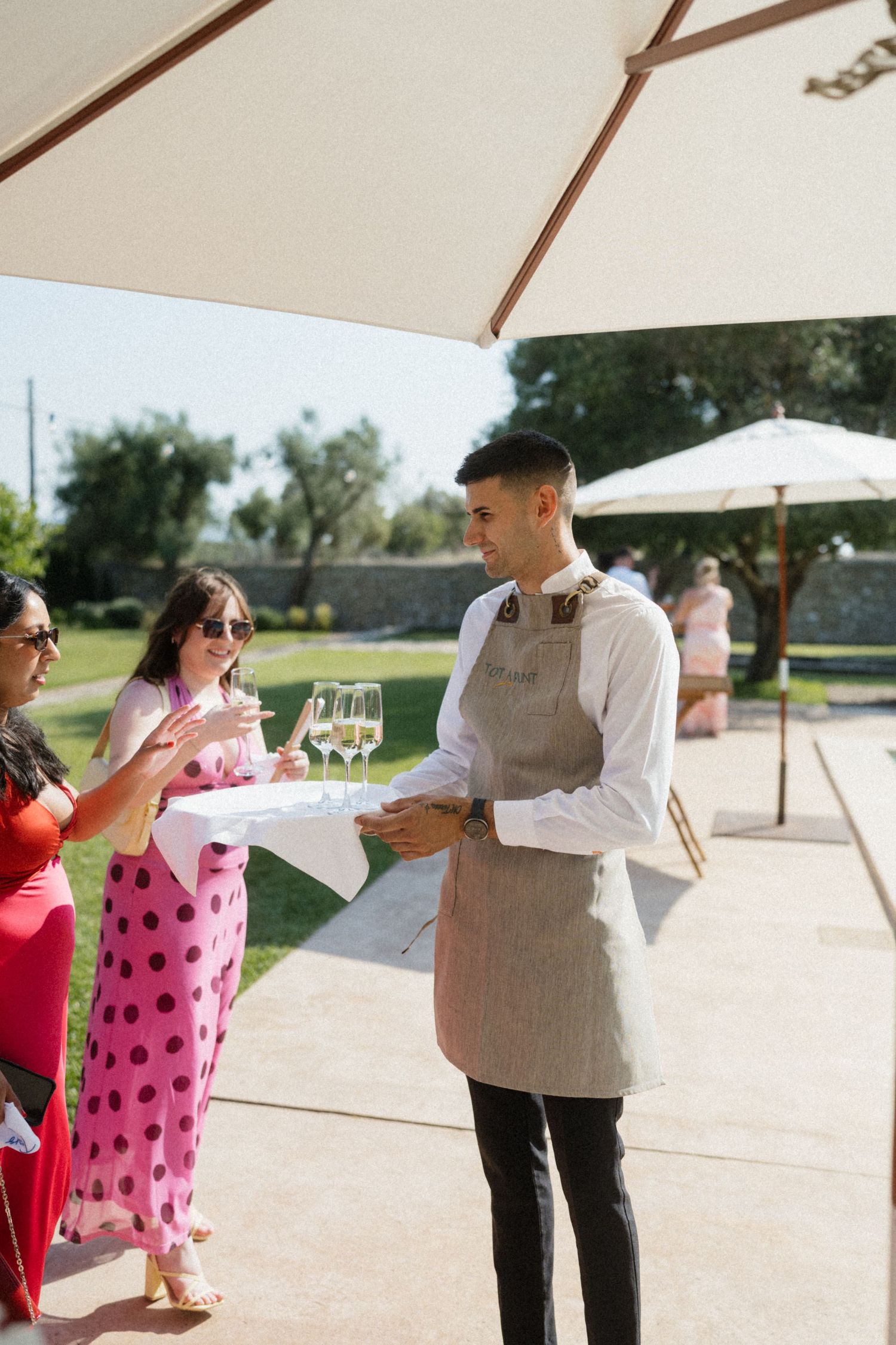 A waiter in a linen apron serves champagne to elegantly dressed guests at an outdoor event on a sunny day.