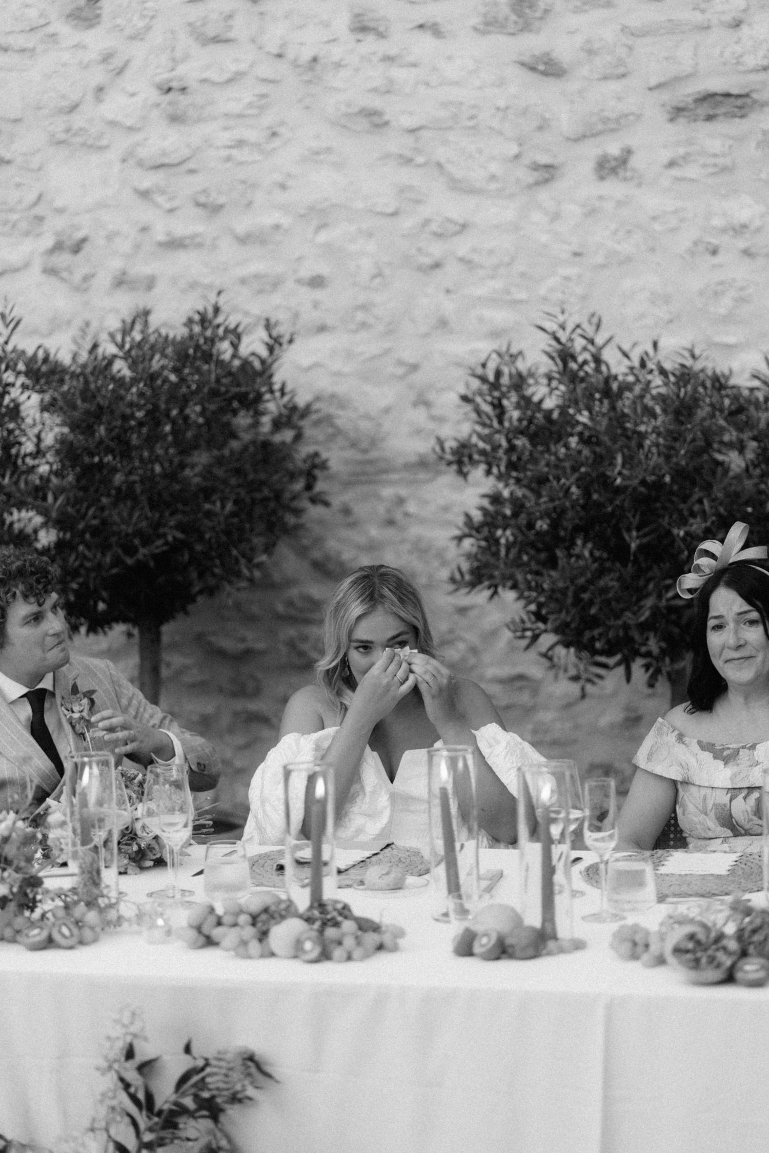 Black and white photo of an emotional bride wiping tears at wedding reception table with candles and fruit centerpieces.
