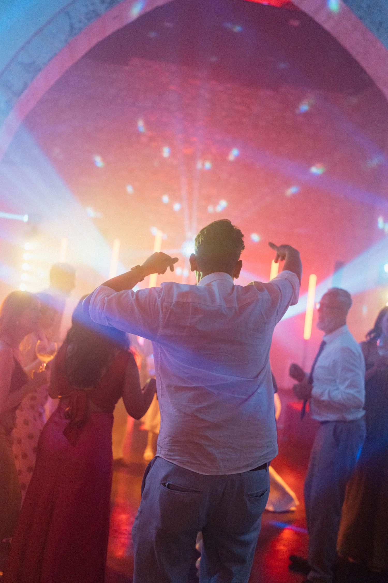 Wedding guests dancing energetically on the dance floor under vibrant colorful lights at a reception.