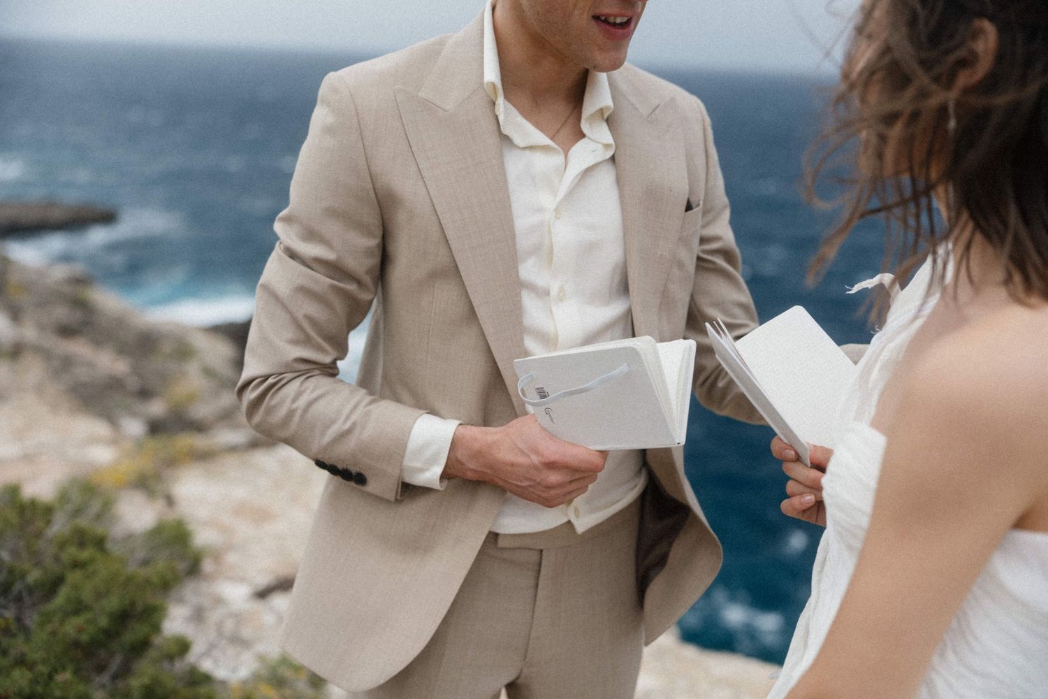 Couple exchanging vows outdoors on a cliff by the ocean, groom in beige suit holding white booklets.