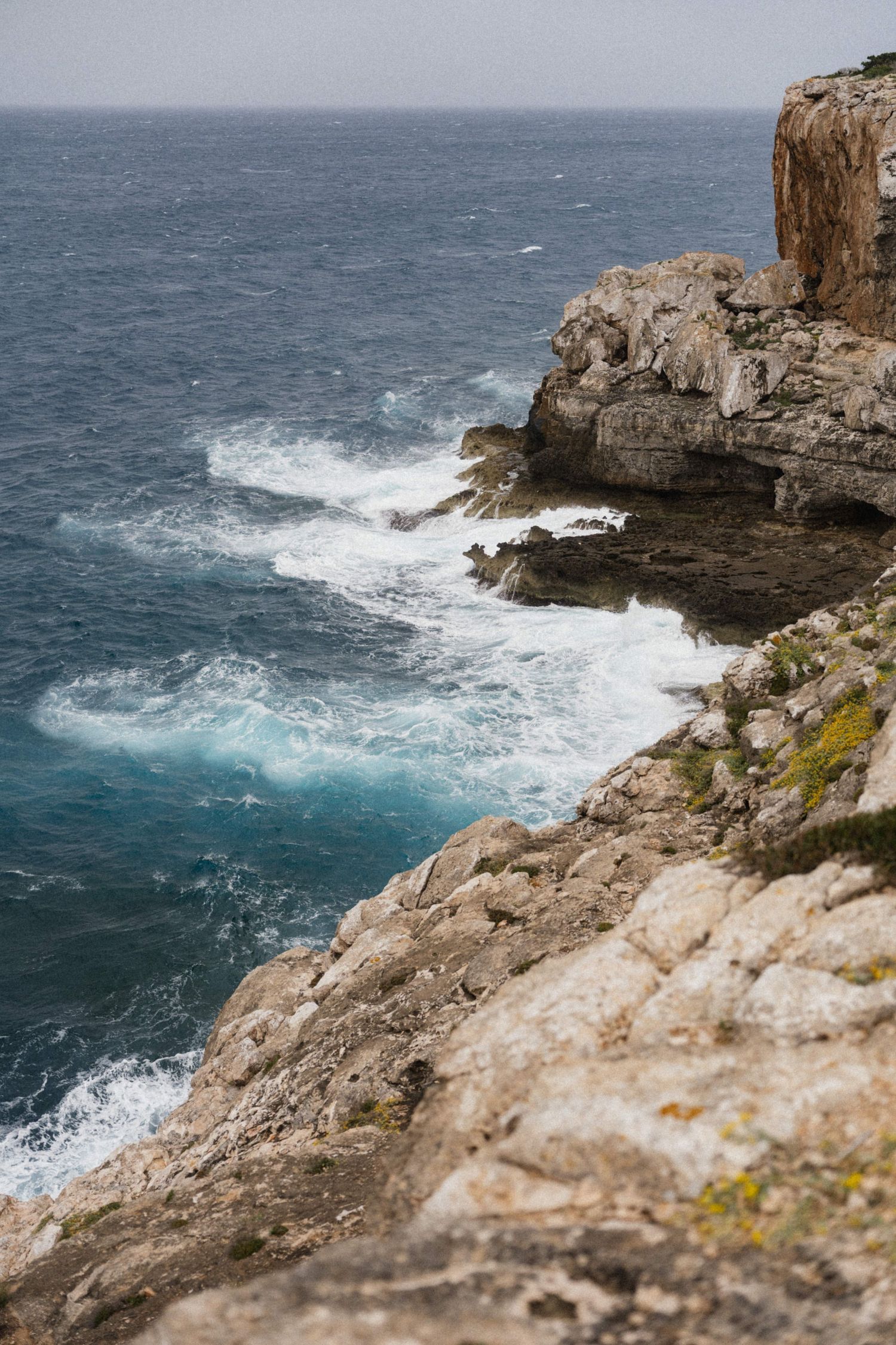 Dramatic rocky coastal cliffs with turquoise ocean waves crashing below on an overcast day.