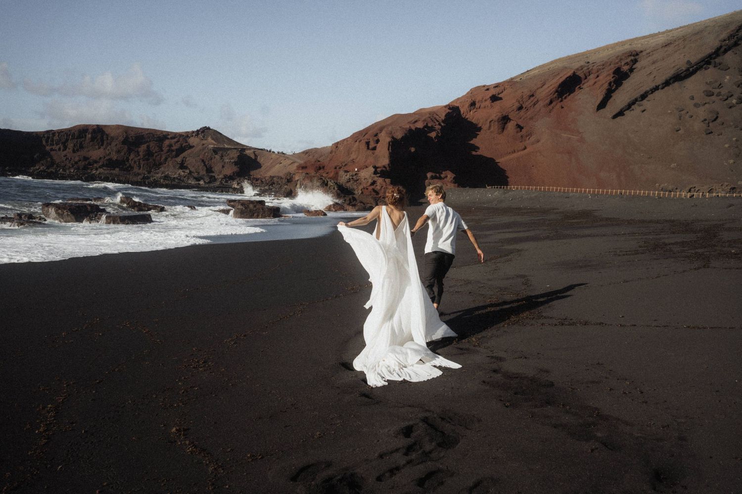Bride and groom running along black sand beach with crashing waves and red cliffs.
