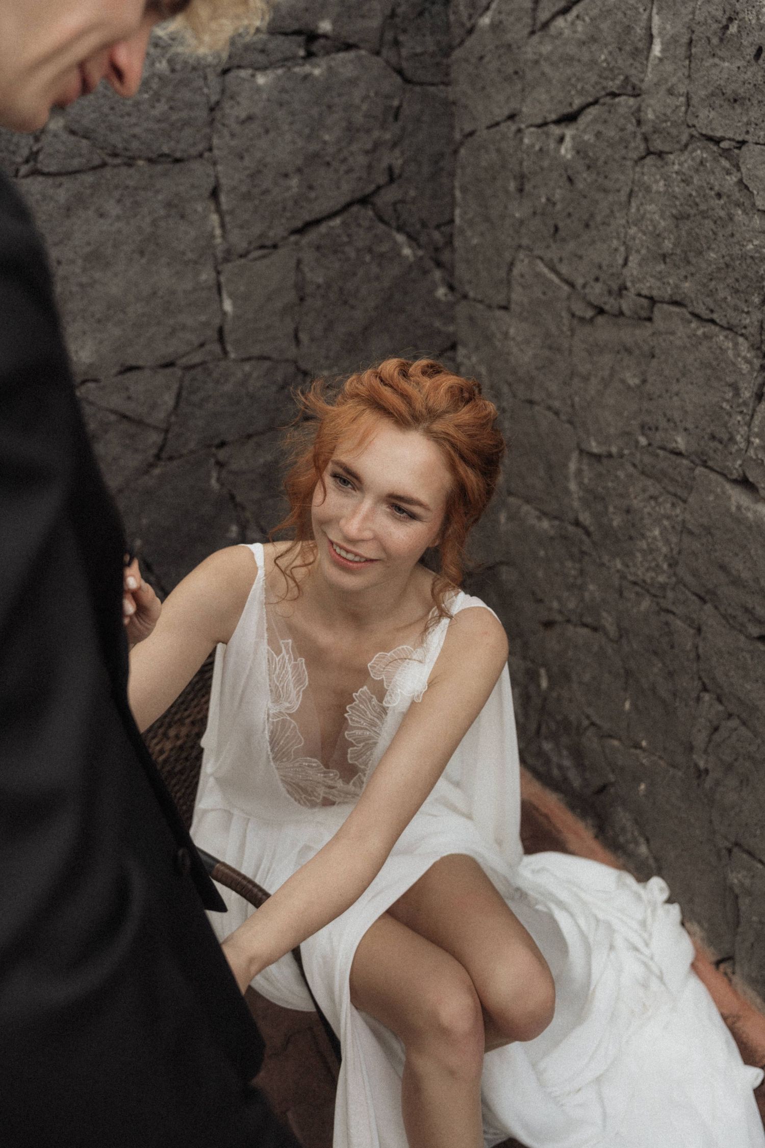 Smiling redhead bride in white lace wedding dress sitting against dark stone wall.