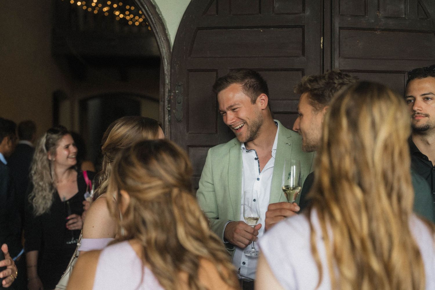 Smiling man in sage green blazer socializing with guests at elegant wedding reception.