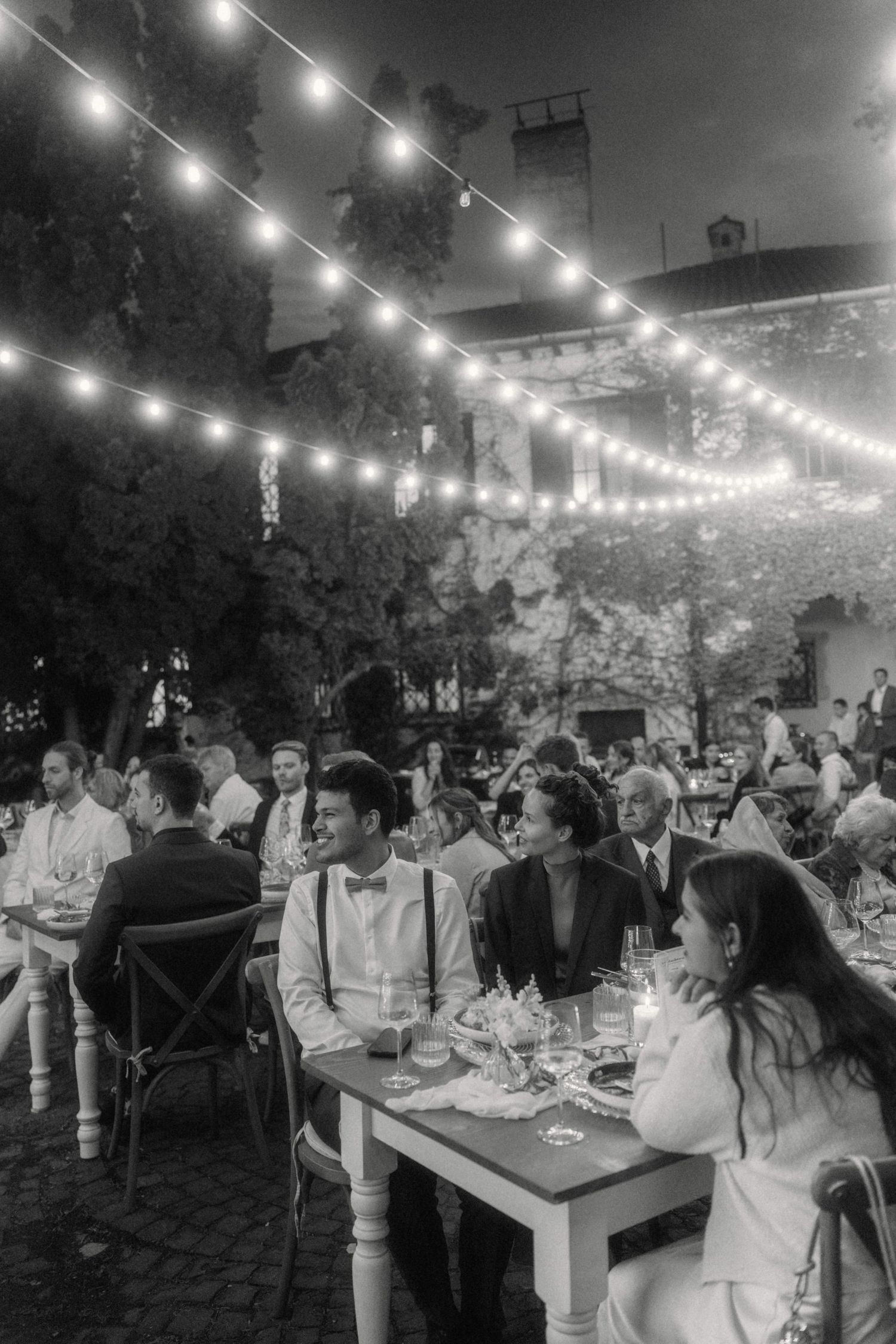 Black and white evening wedding reception with guests dining under glowing string lights outdoors.