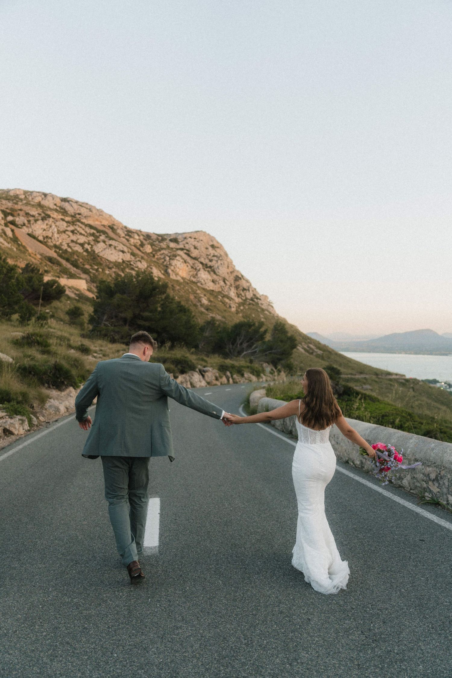 Bride and groom walk hand-in-hand down a scenic coastal road surrounded by mountains at sunset.