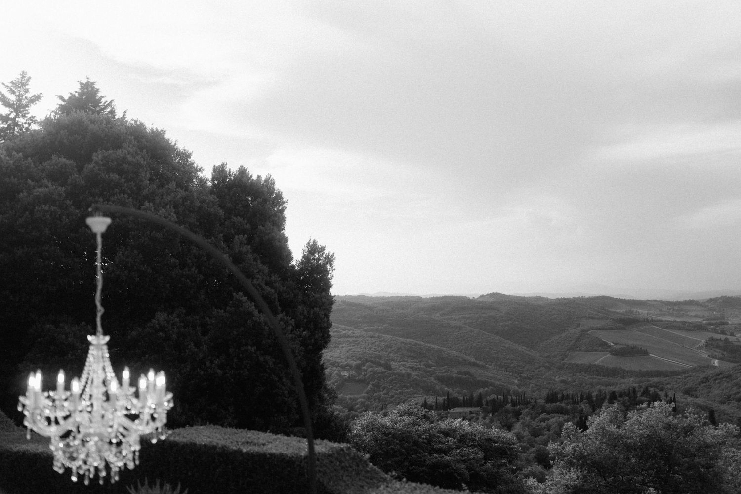 Elegant crystal chandelier hanging outdoors with a scenic Tuscan countryside view in black and white.