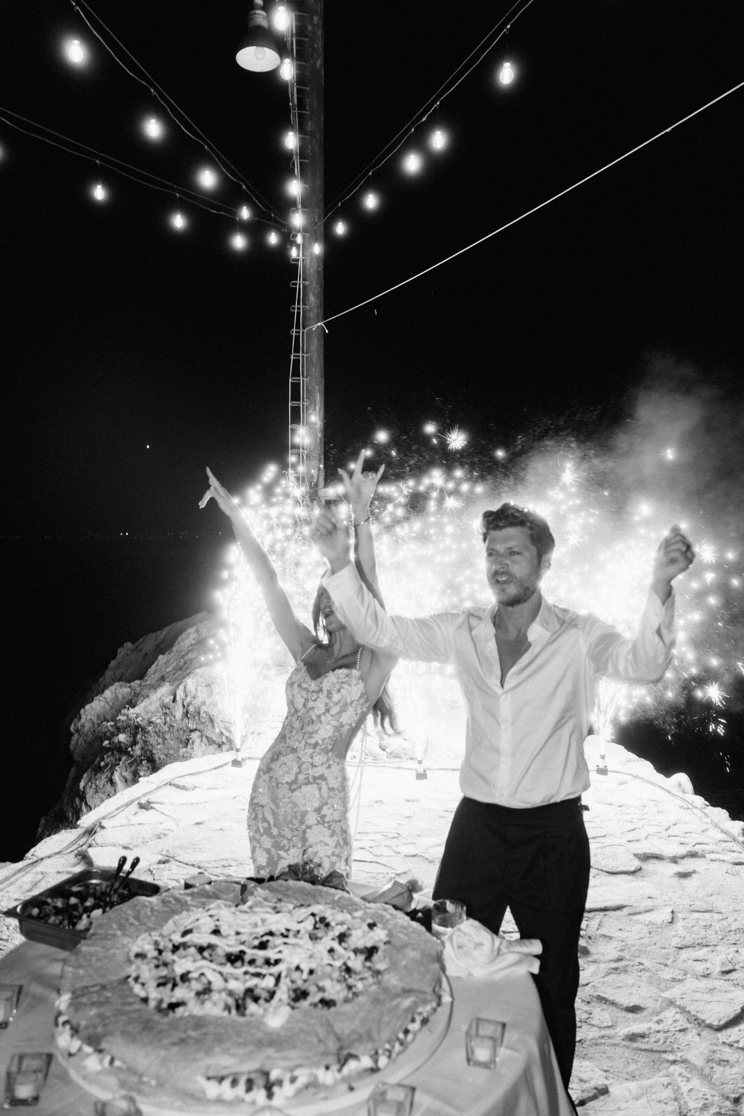 Black and white photo of bride and groom celebrating with fireworks and wedding cake at night outdoor reception.