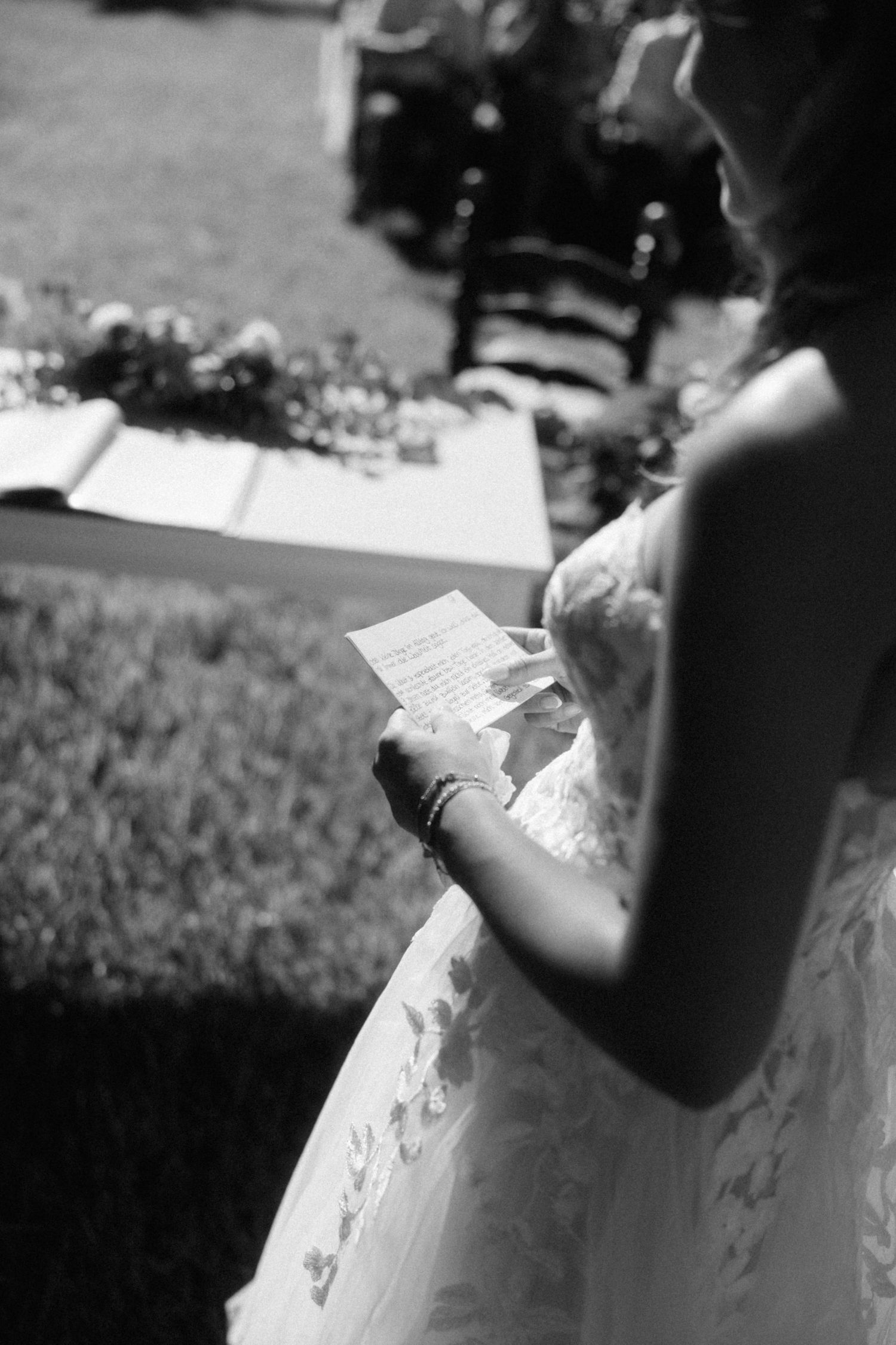 Black and white photo of a bride in a floral gown reading her wedding vows from a card.