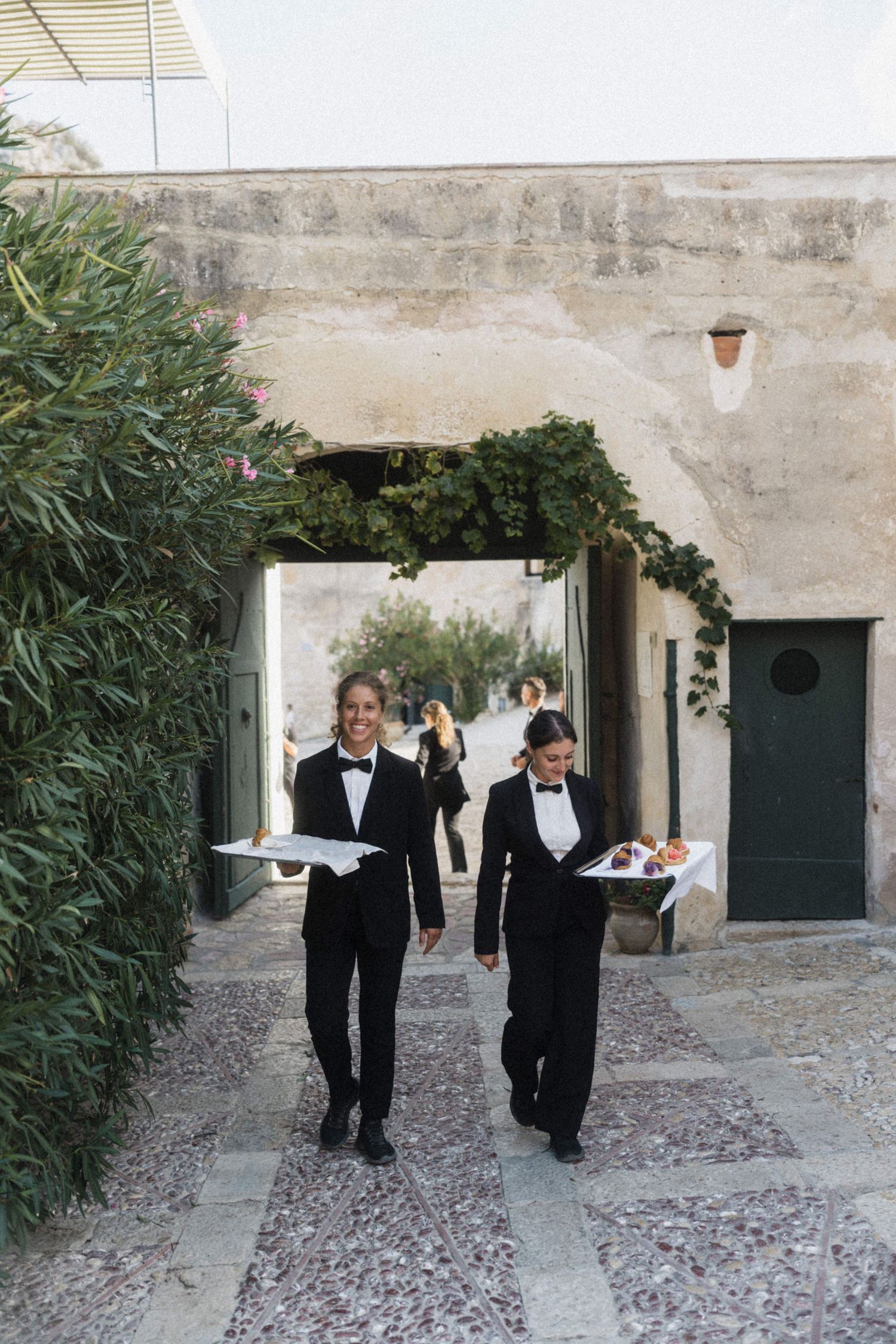 Two smiling waiters in black tuxedos carrying silver trays through a vine-covered archway.