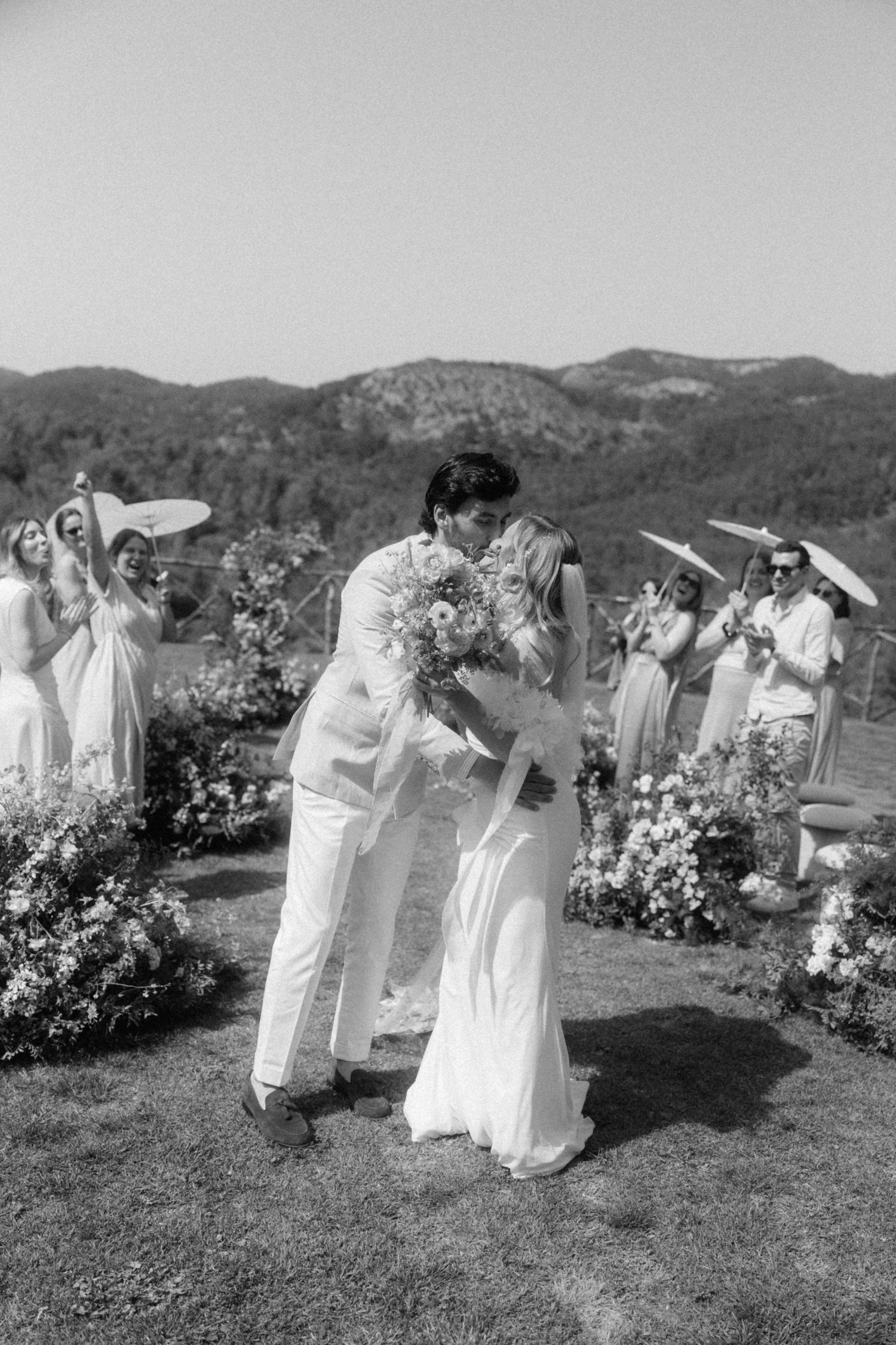 Black and white photo of newlyweds sharing first kiss at outdoor wedding ceremony with mountain backdrop.