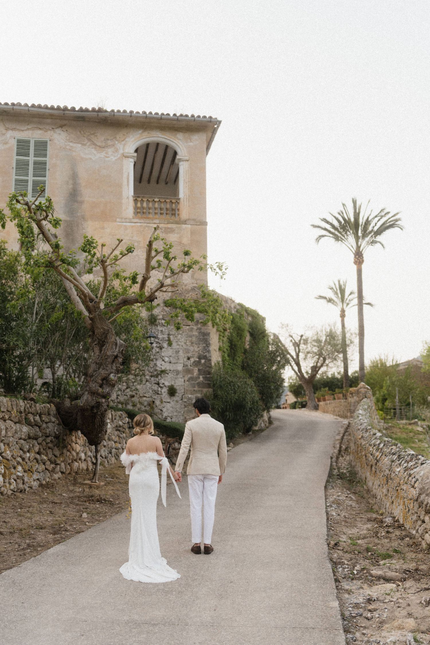 Bride and groom walk hand in hand along a stone path past a rustic villa with palm trees in Mallorca.