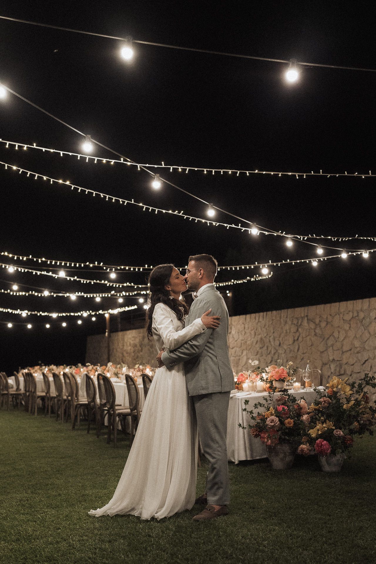 Bride and groom share a romantic kiss under string lights at an outdoor evening wedding reception with floral arrangements.