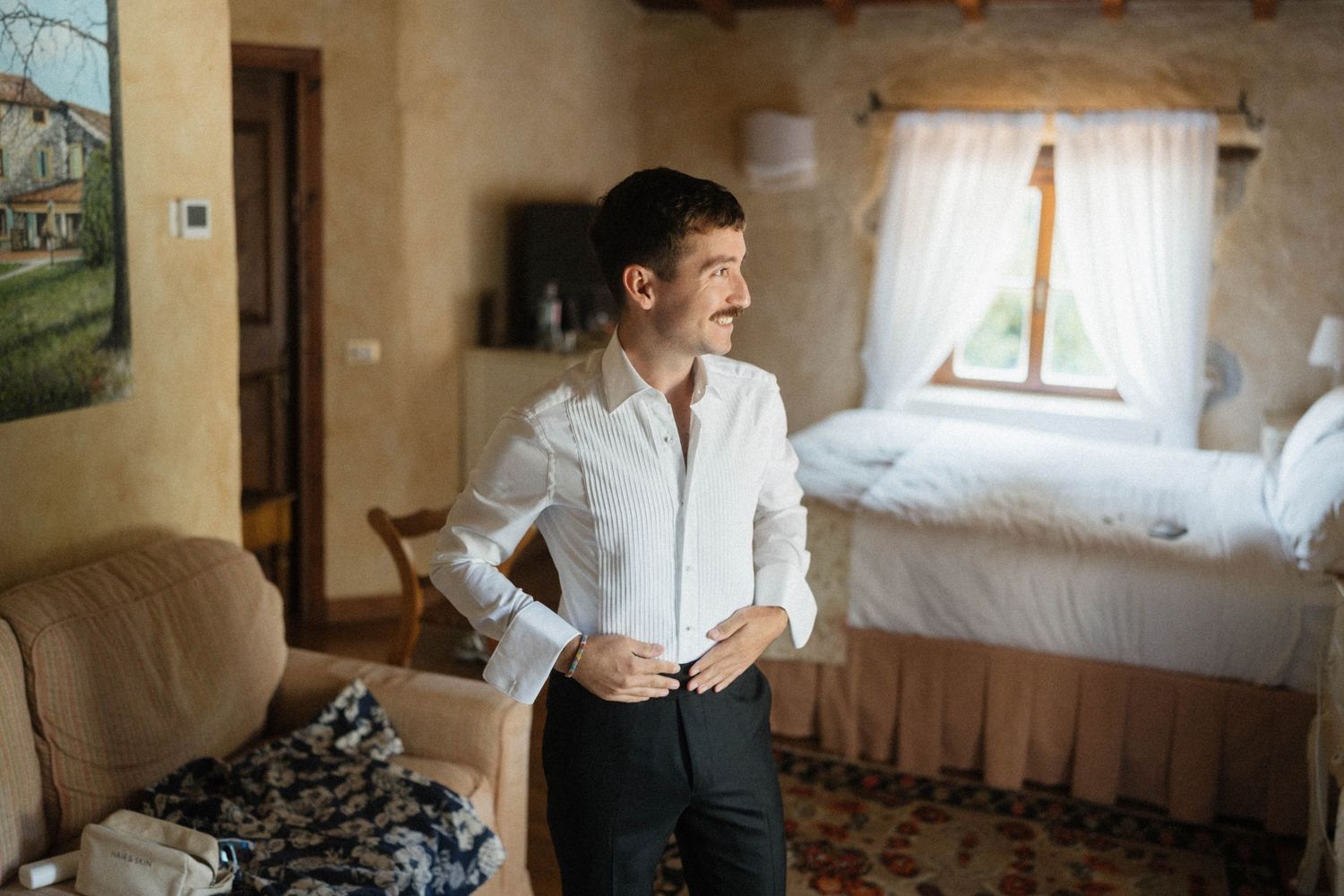 Groom buttoning his white dress shirt while getting ready in a rustic stone villa bedroom.