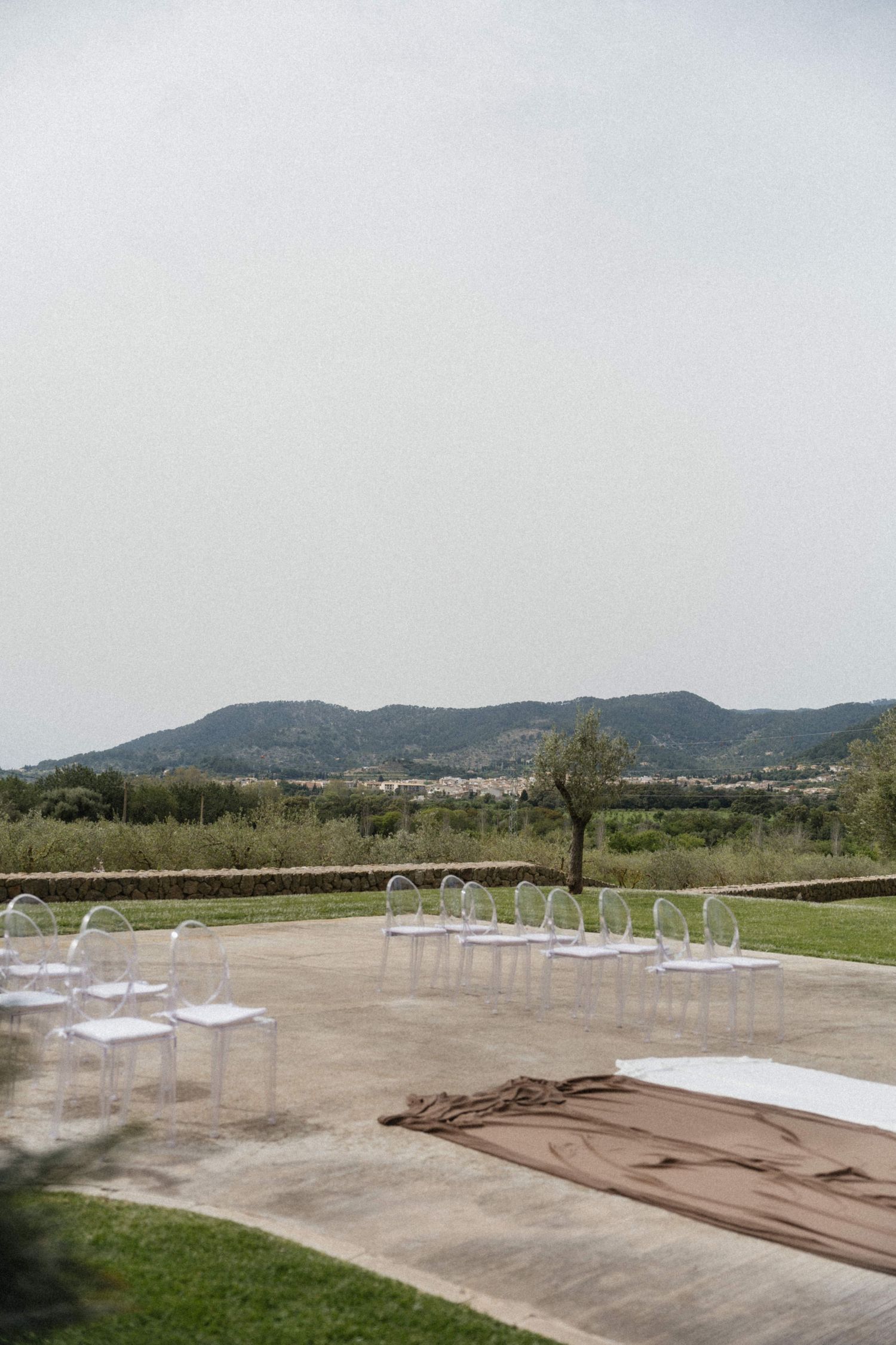 Elegant outdoor wedding ceremony setup with ghost chairs and brown fabric aisle runner with mountain views.