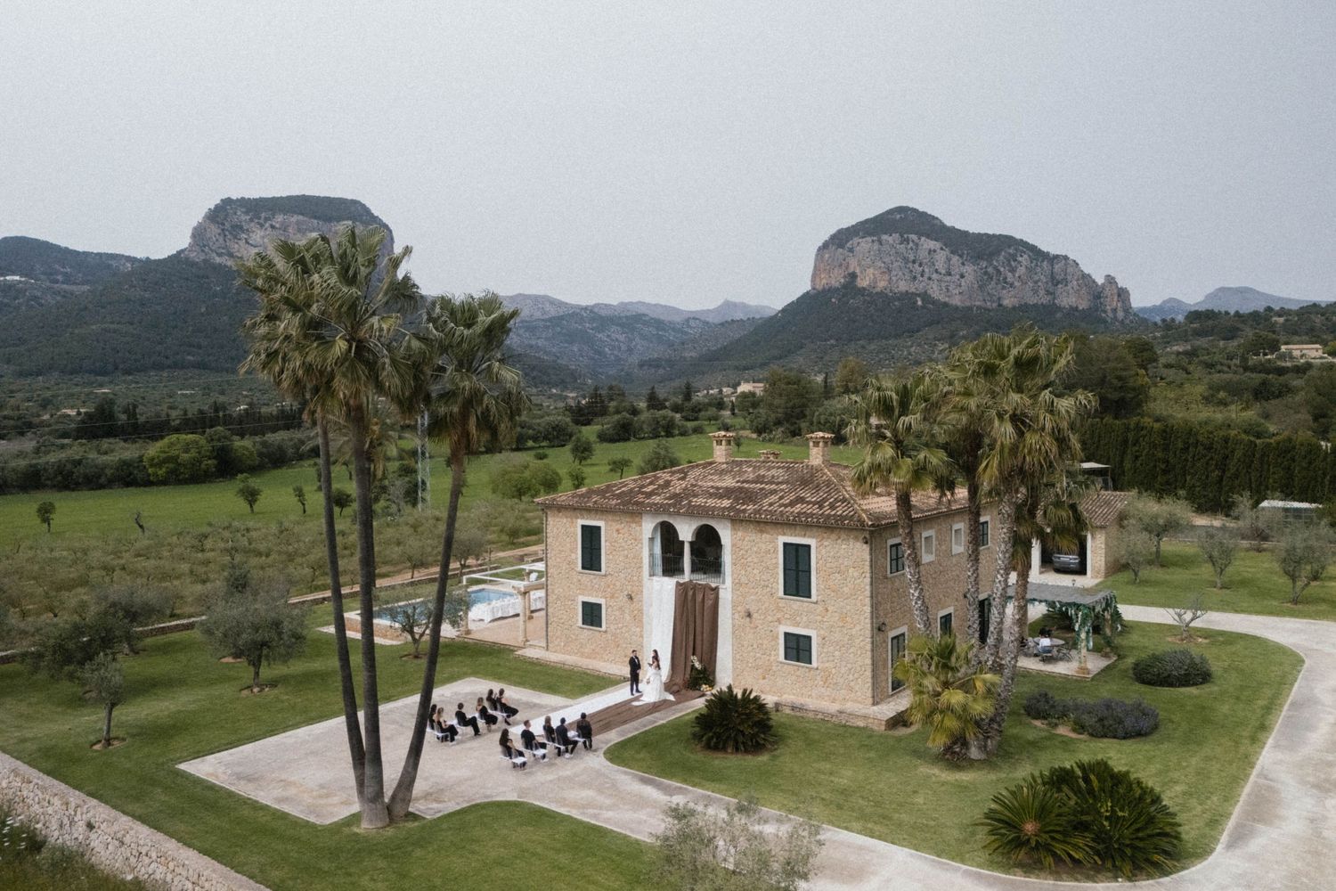 Aerial view of rustic stone finca surrounded by palm trees with dramatic mountain backdrop in Mallorca, Spain.