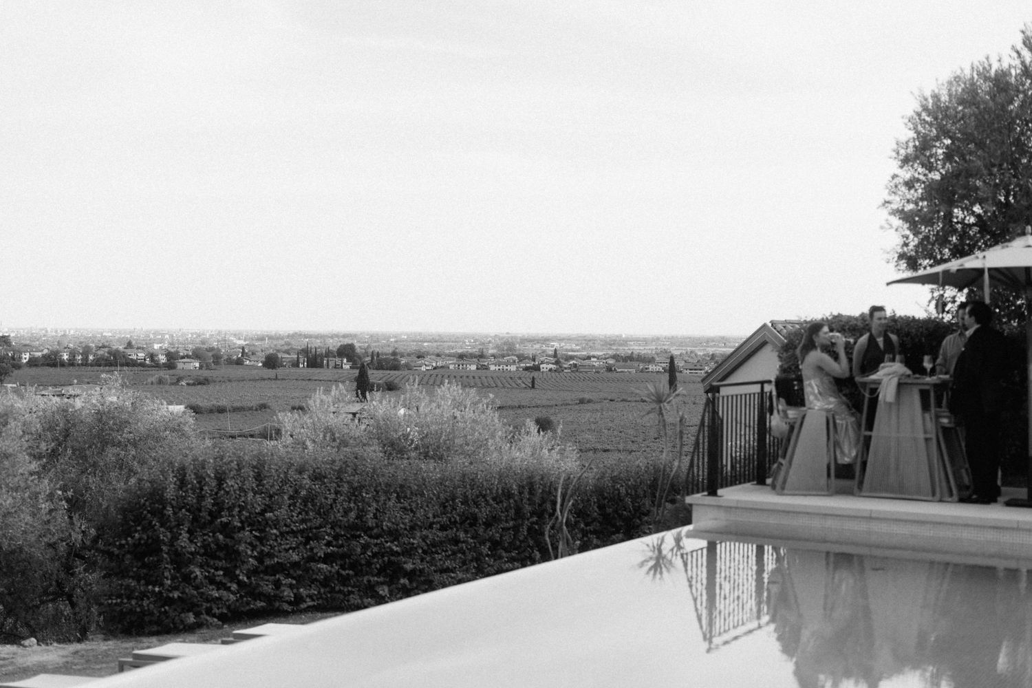 Black and white panoramic view of wedding guests gathered by a pool overlooking a scenic countryside landscape.