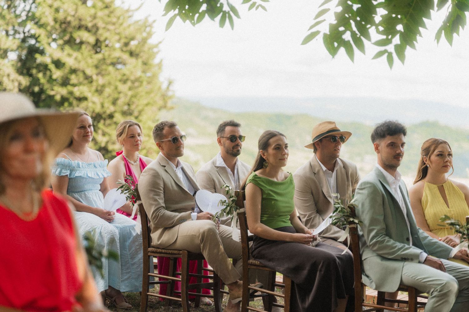 Guests seated outdoors at a scenic hillside wedding ceremony, dressed in elegant summer attire.