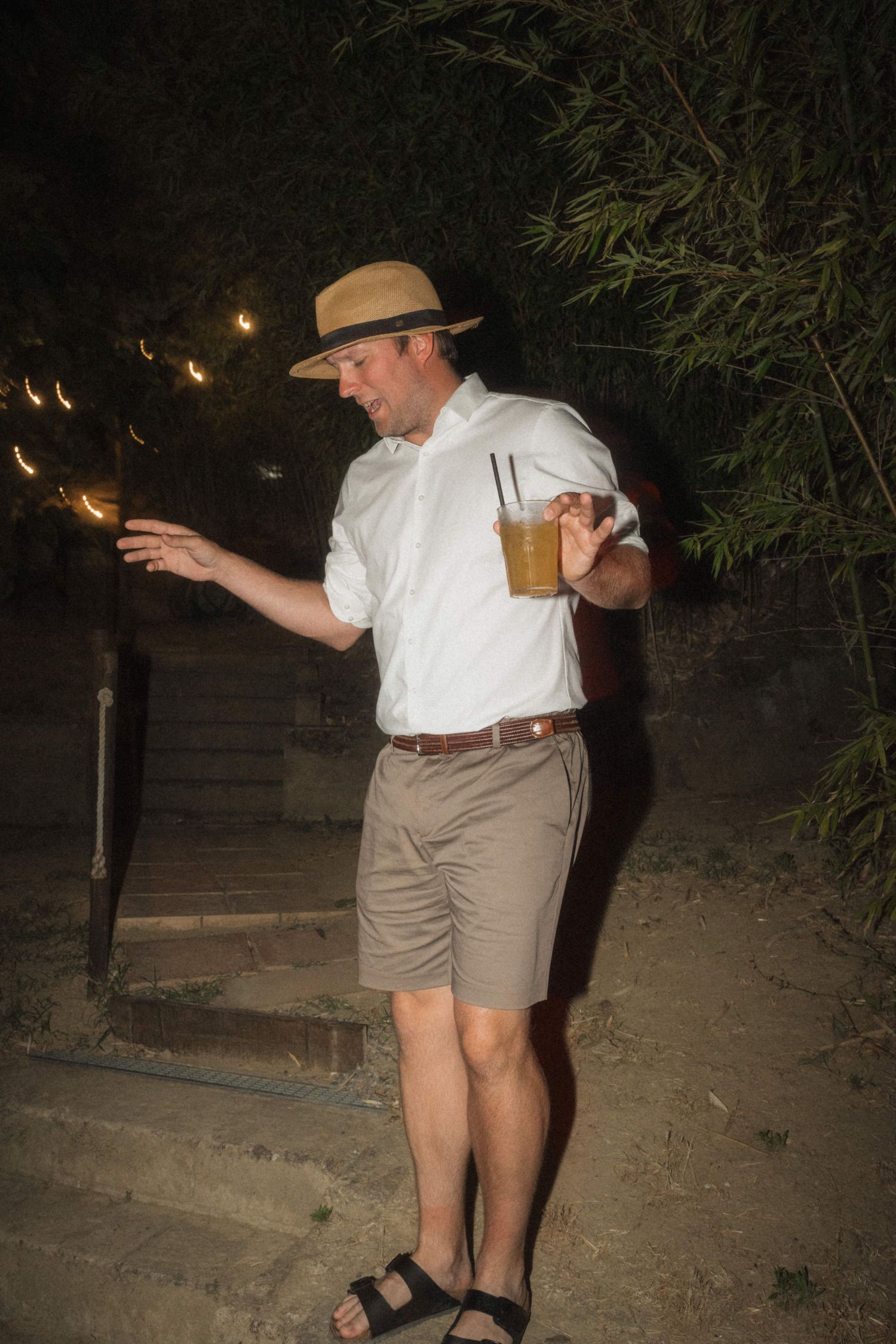 Man in straw hat and white shirt holding sparkler and drink at an outdoor evening wedding reception.