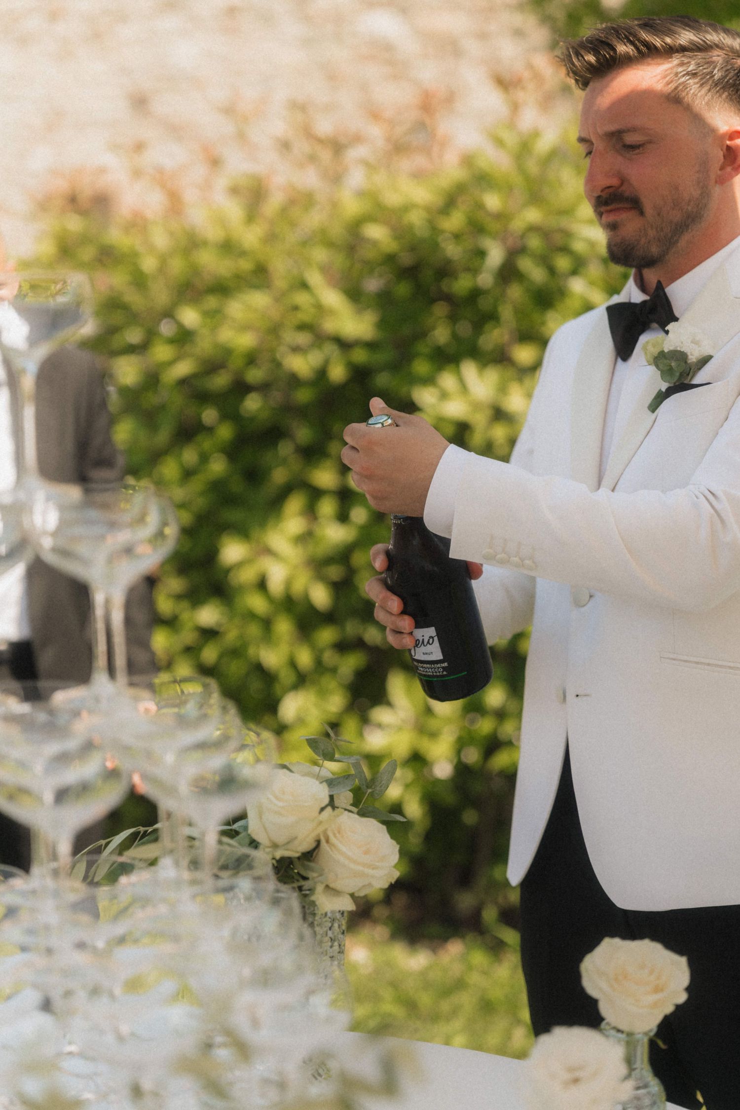 Groom in white tuxedo opening champagne bottle beside elegant glass tower with white roses at outdoor wedding.